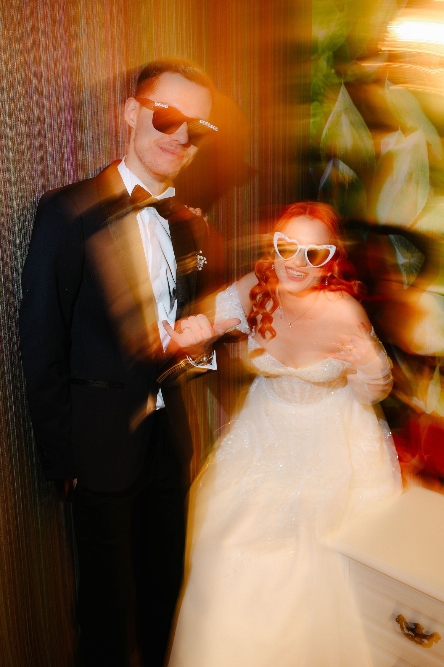 A bride in white dress and groom in black tuxedo wearing novelty sunglasses pose together in a fun blurred wedding photo.