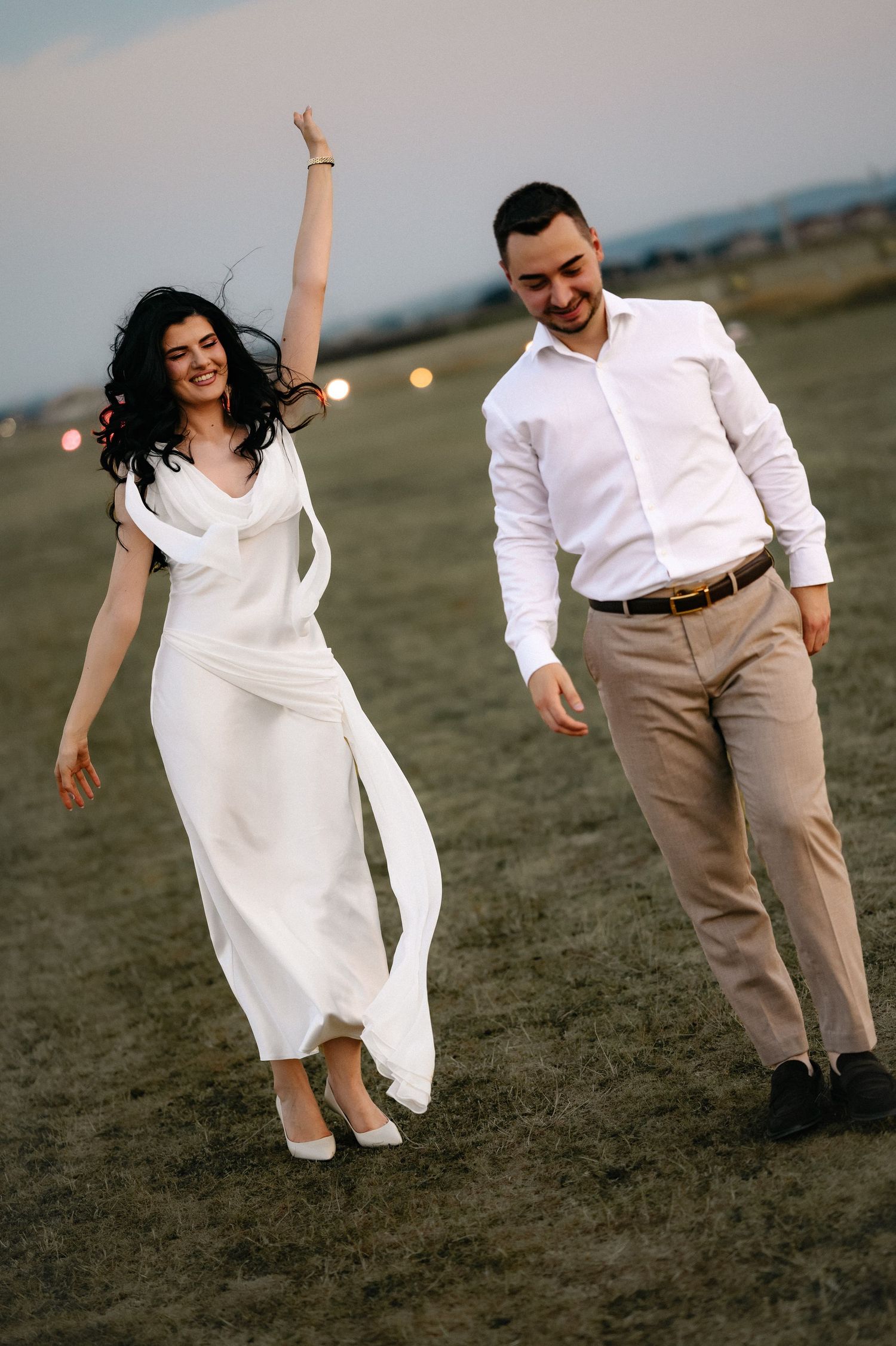 Joyful bride in white dress raises arm while groom in white shirt smiles, walking together in field at dusk.