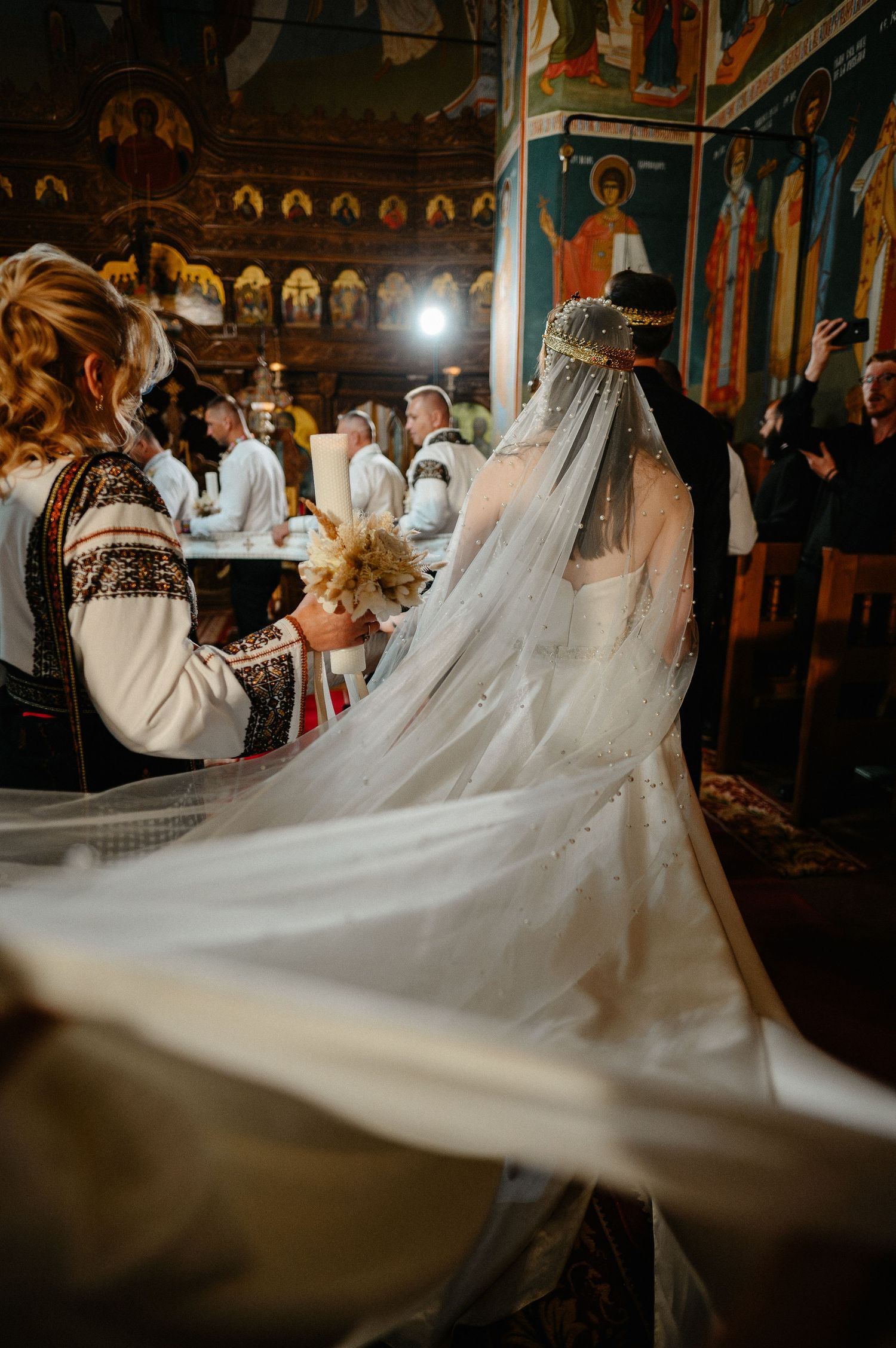 Bride in pearl veil with long train stands at Orthodox church altar during wedding ceremony.