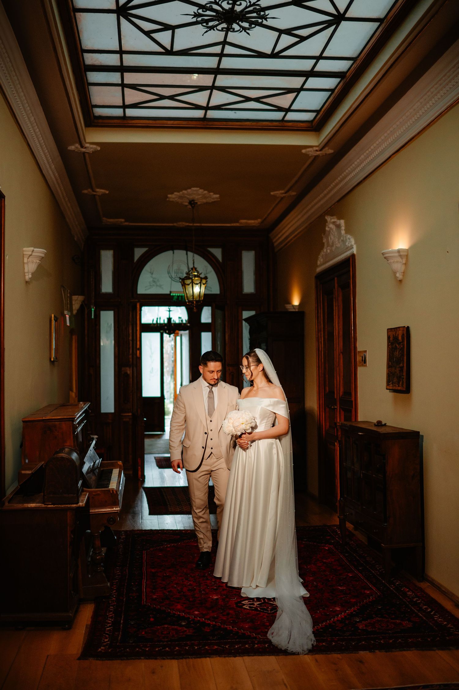 Bride and groom walking together through elegant vintage hallway with decorative glass ceiling.