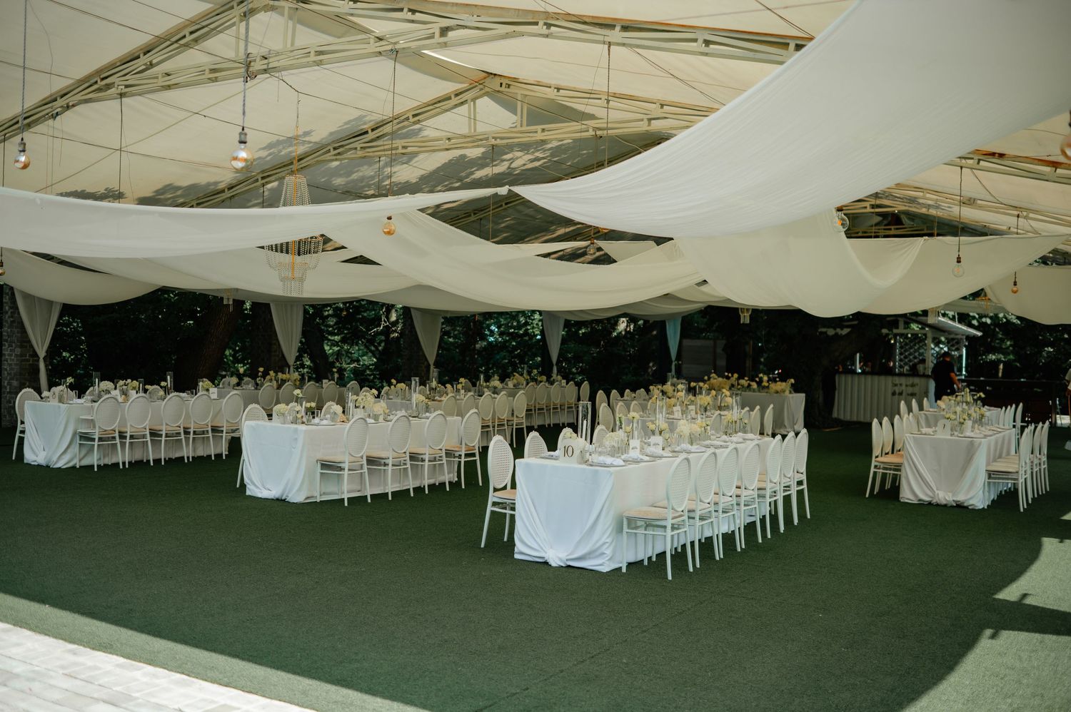 Elegant outdoor wedding reception under transparent tent with white draped ceiling and floral centerpieces.