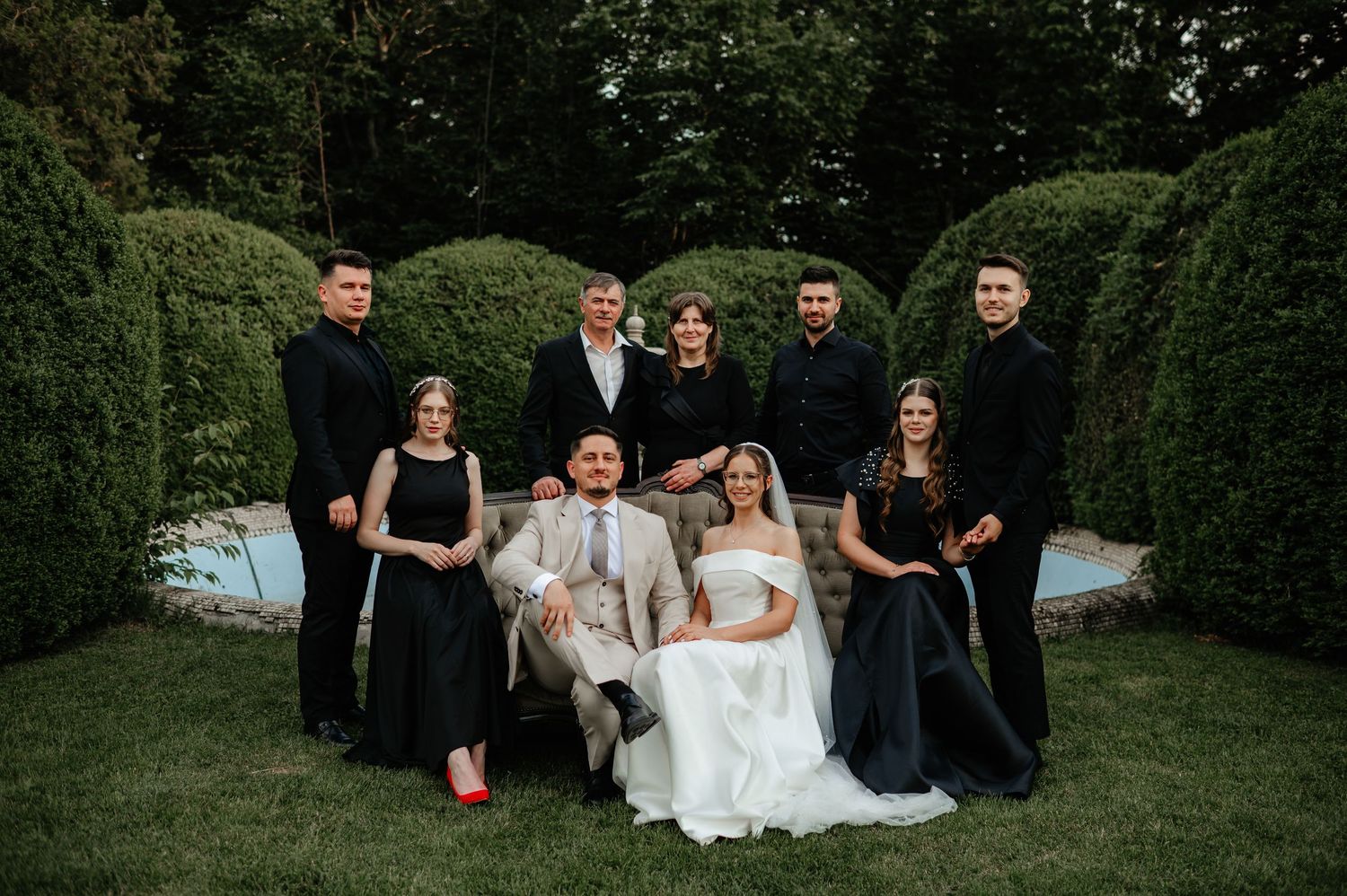 Wedding couple posing with family group in formal attire among manicured garden hedges.