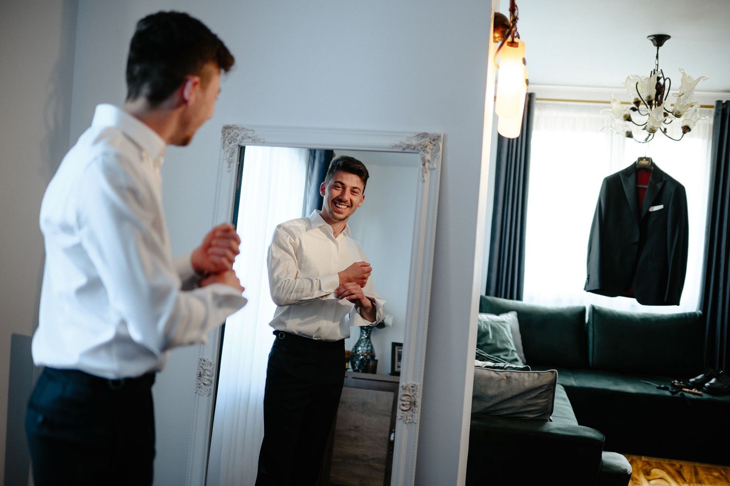 Groom preparing for wedding day, adjusting cufflinks while smiling at mirror reflection.