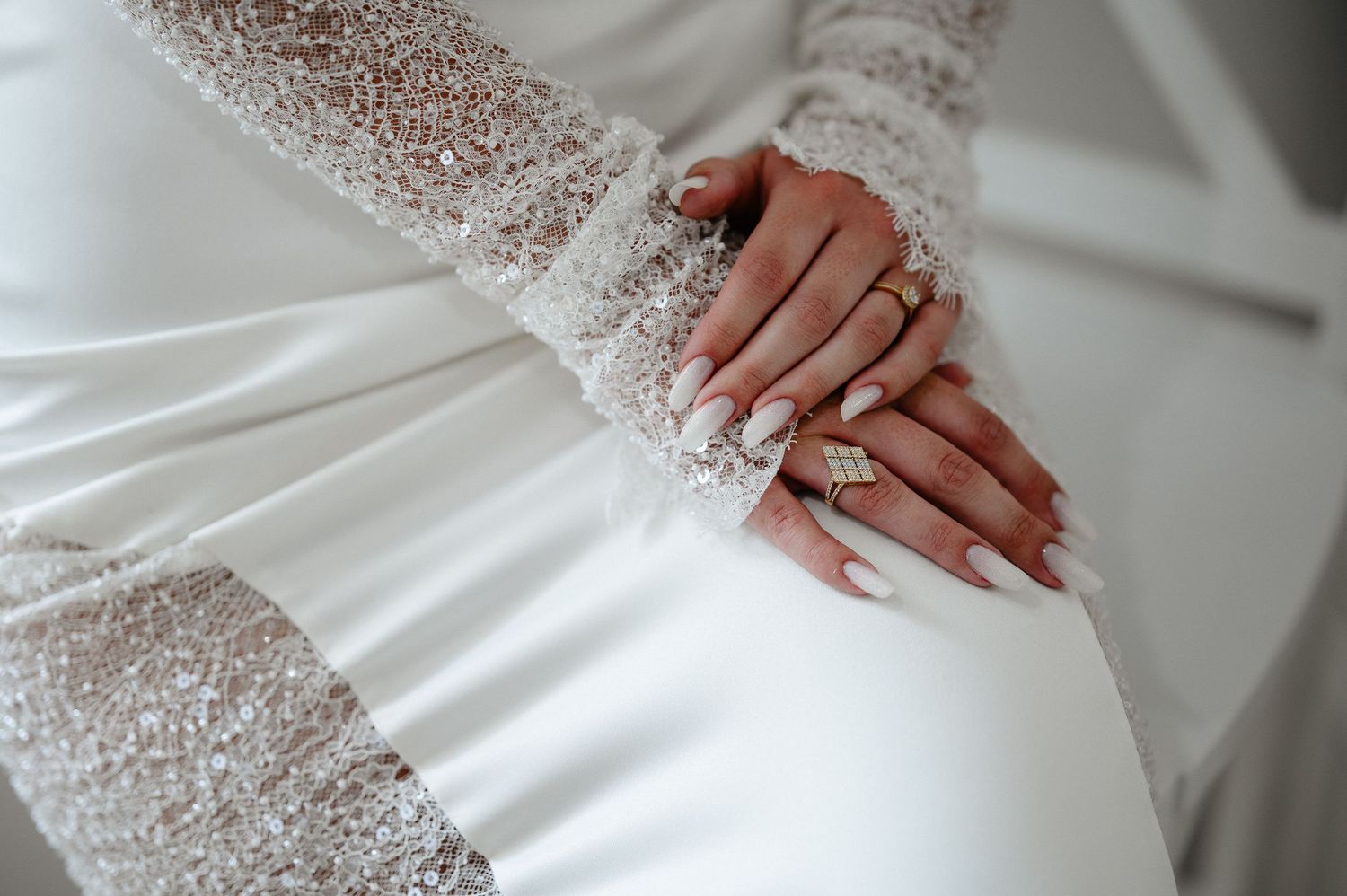 Close-up of bride's hands with gold rings and white nails resting on lace wedding dress.