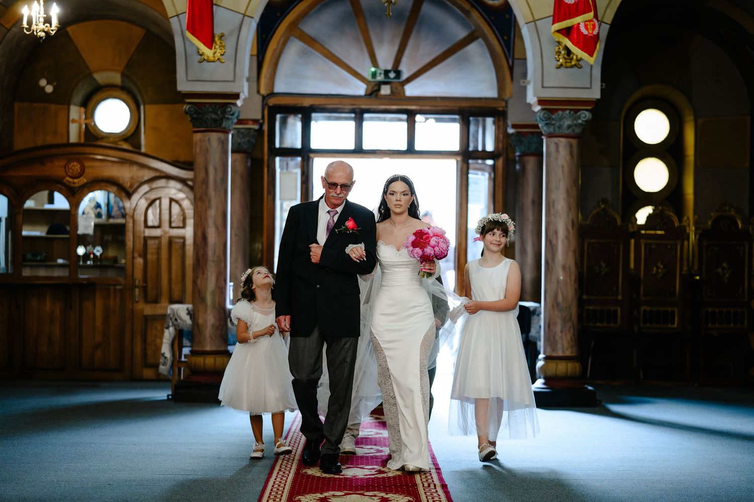 Bride in white gown walks down church aisle with father and two flower girls at wedding ceremony.