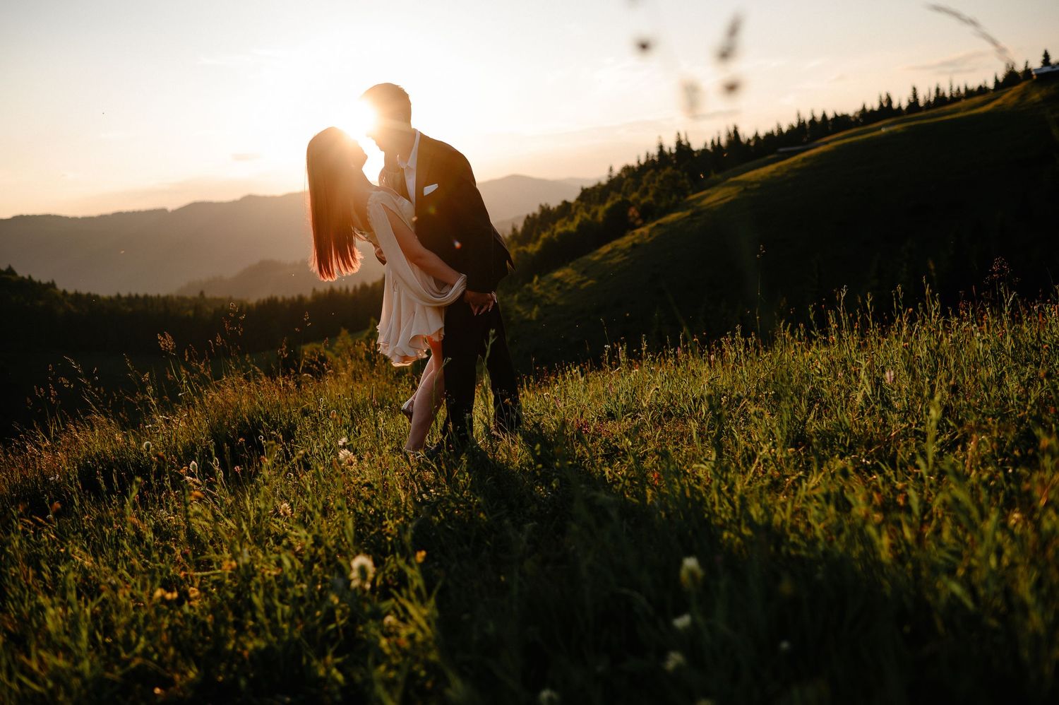 A couple shares a romantic kiss on a sunlit hillside with mountains and wildflowers at sunset.
