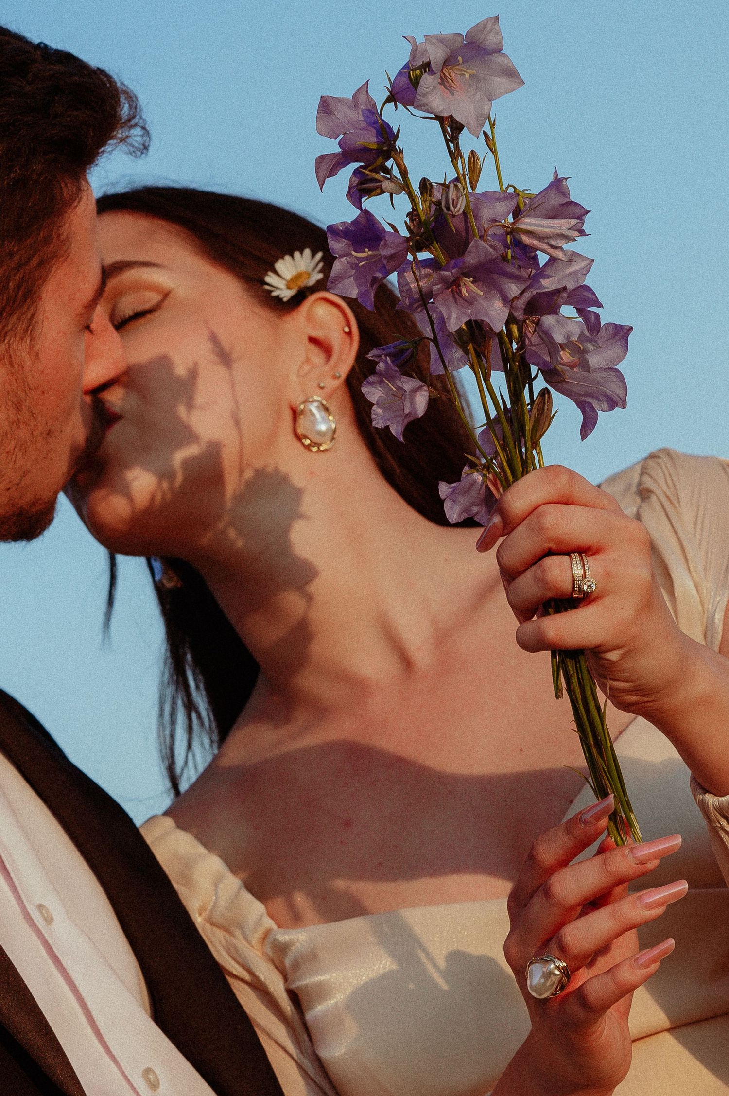 Couple kissing with bride holding purple wildflowers and daisy hair accessory against a clear blue sky.