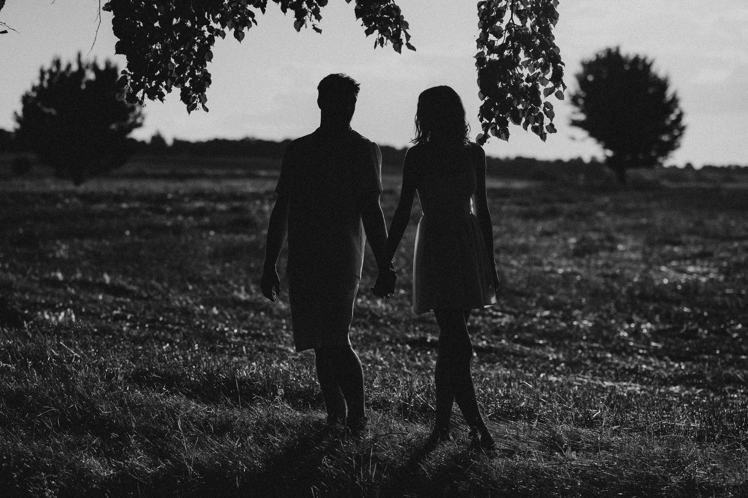 Silhouette of couple holding hands walking through open field at dusk.