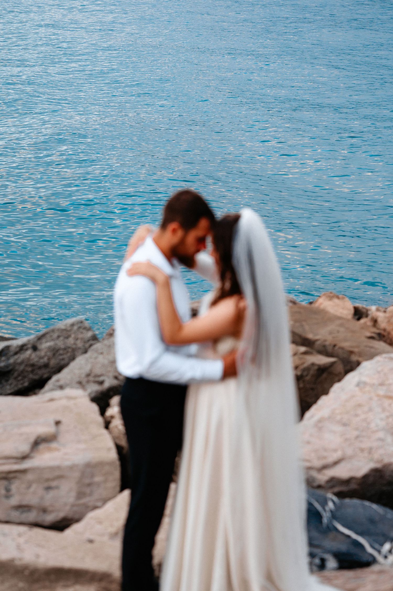 Blurred artistic shot of bride and groom embracing near vivid blue ocean water.