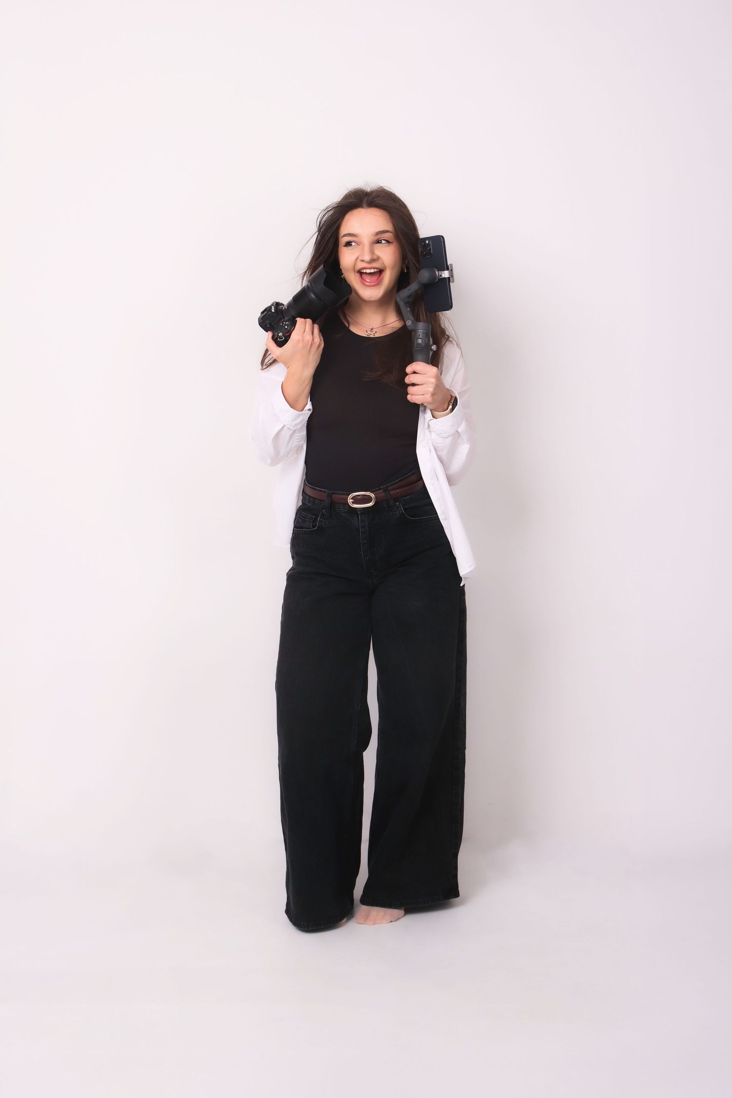 Excited young woman in black and white outfit holding cameras against a white studio background.