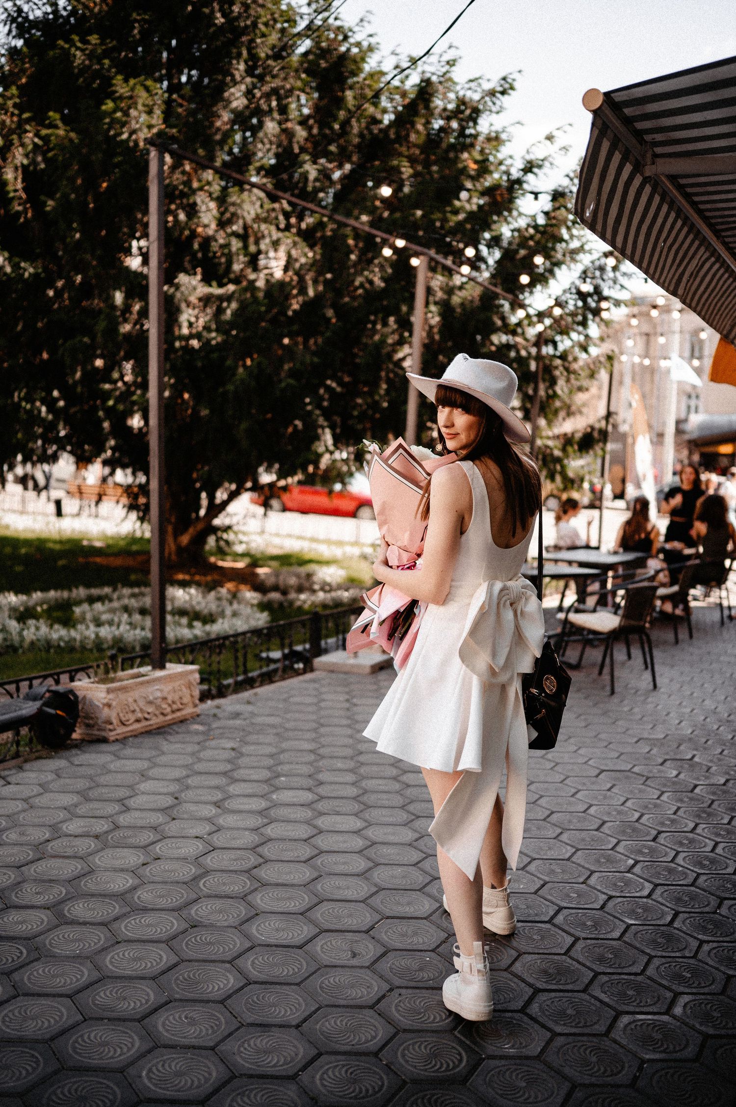 Stylish young woman in white dress and wide-brim hat walking on a tiled urban plaza on a sunny day.