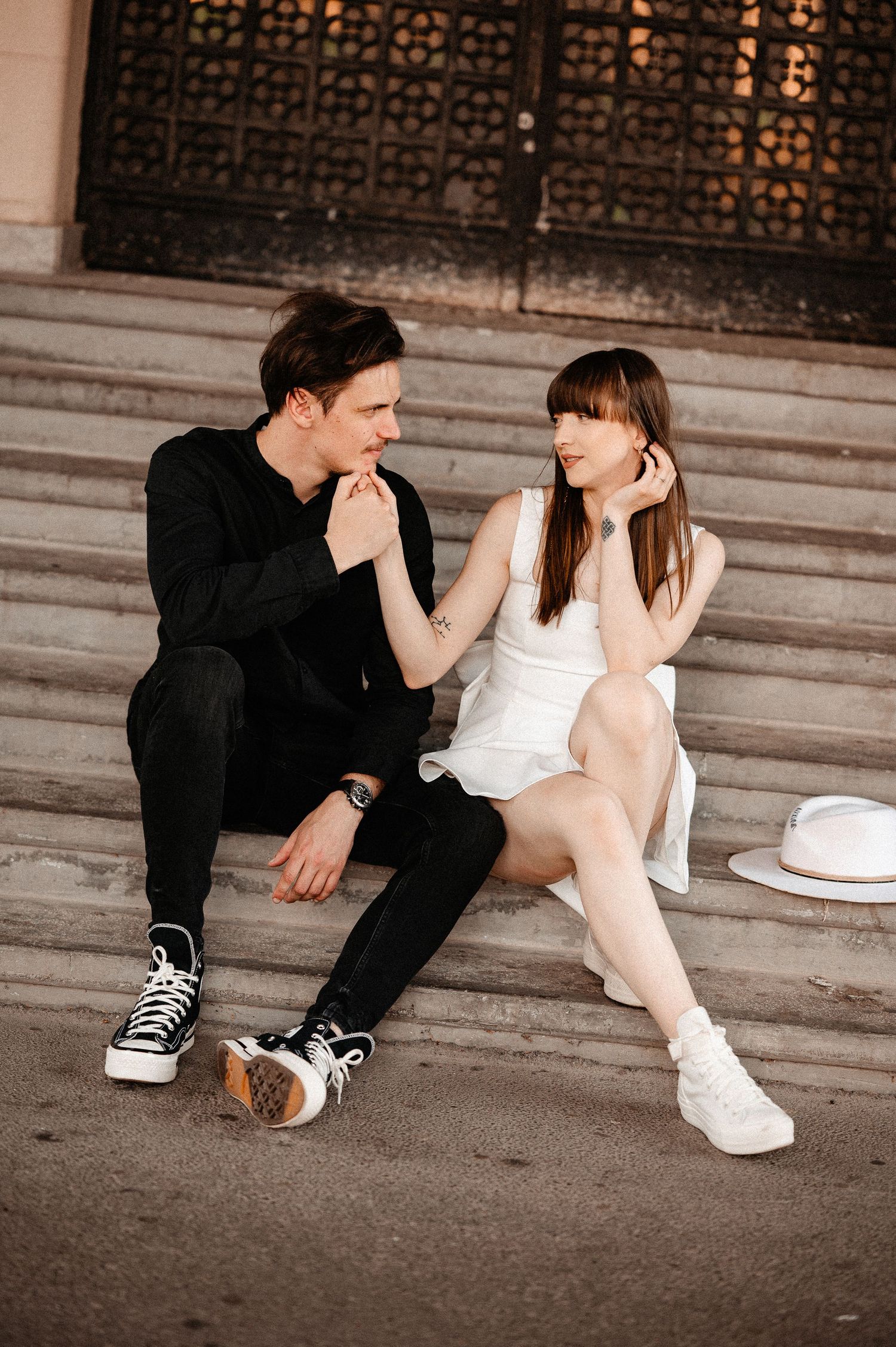 Young couple sitting on stone steps, smiling and holding hands in casual chic outfits.