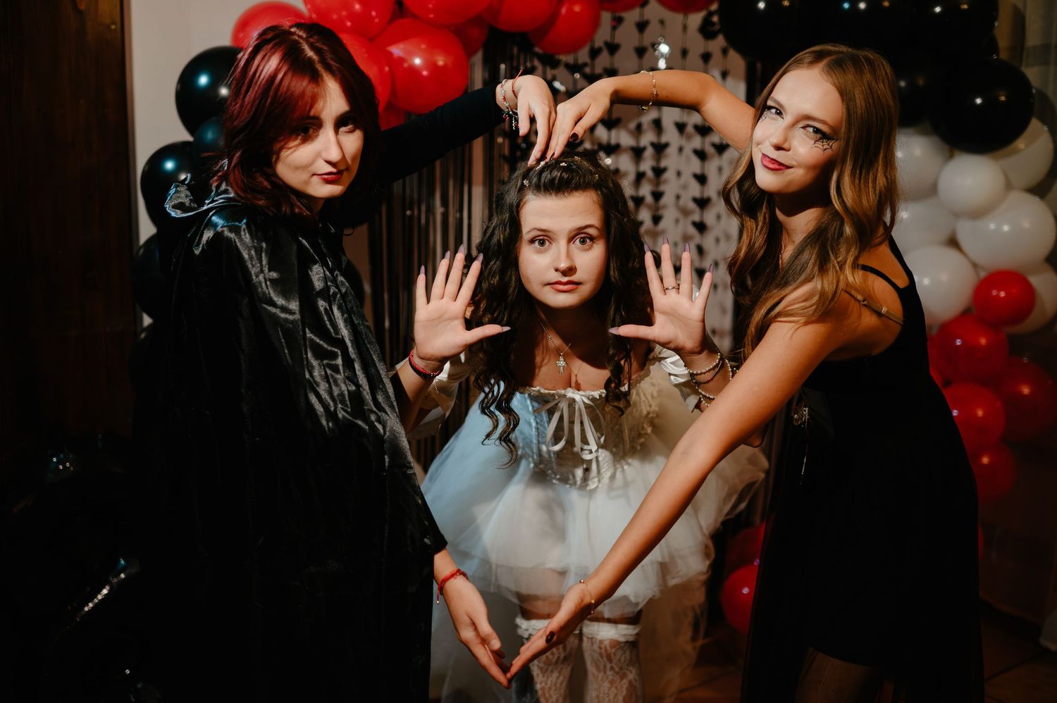 Three women in Halloween costumes pose playfully in front of red, black, and white balloon decorations.