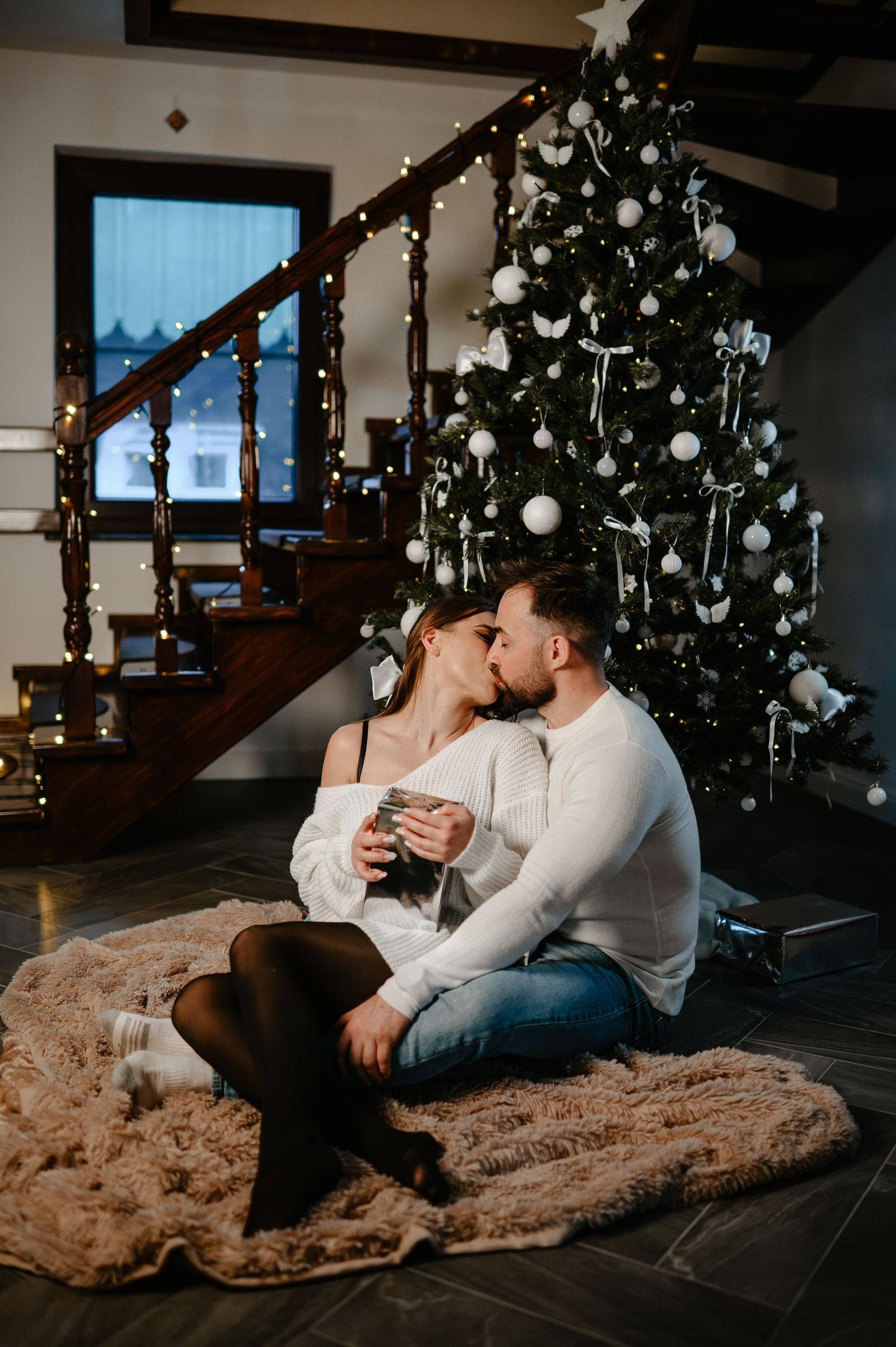 Romantic couple kissing while sitting on a fur rug near a decorated Christmas tree.