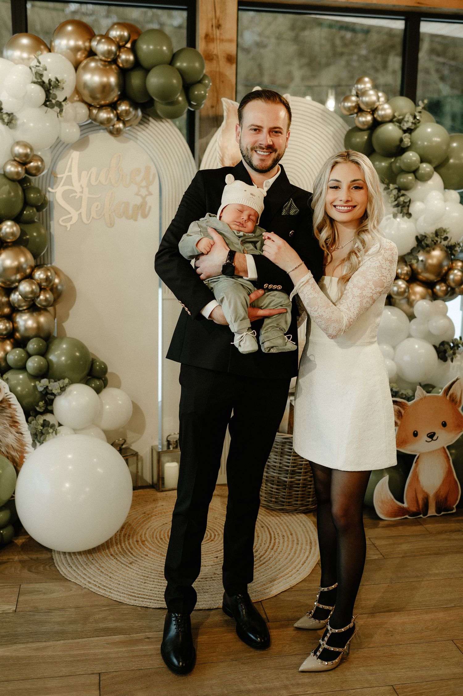 Smiling couple in formal attire holding their newborn at an elegant woodland baby christening celebration with balloon decor.