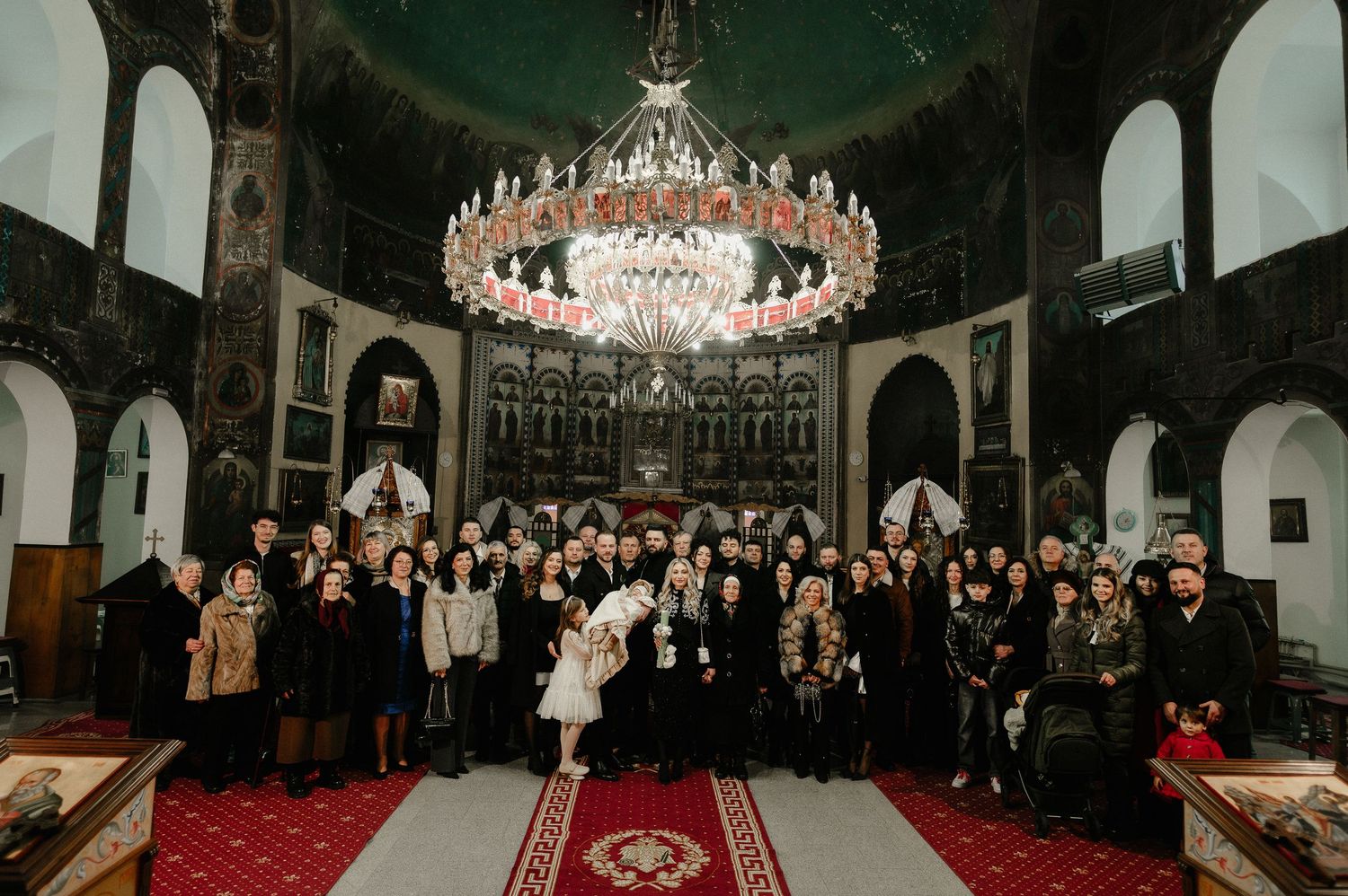 Large group photo inside ornate Orthodox church with chandelier during baptism ceremony.