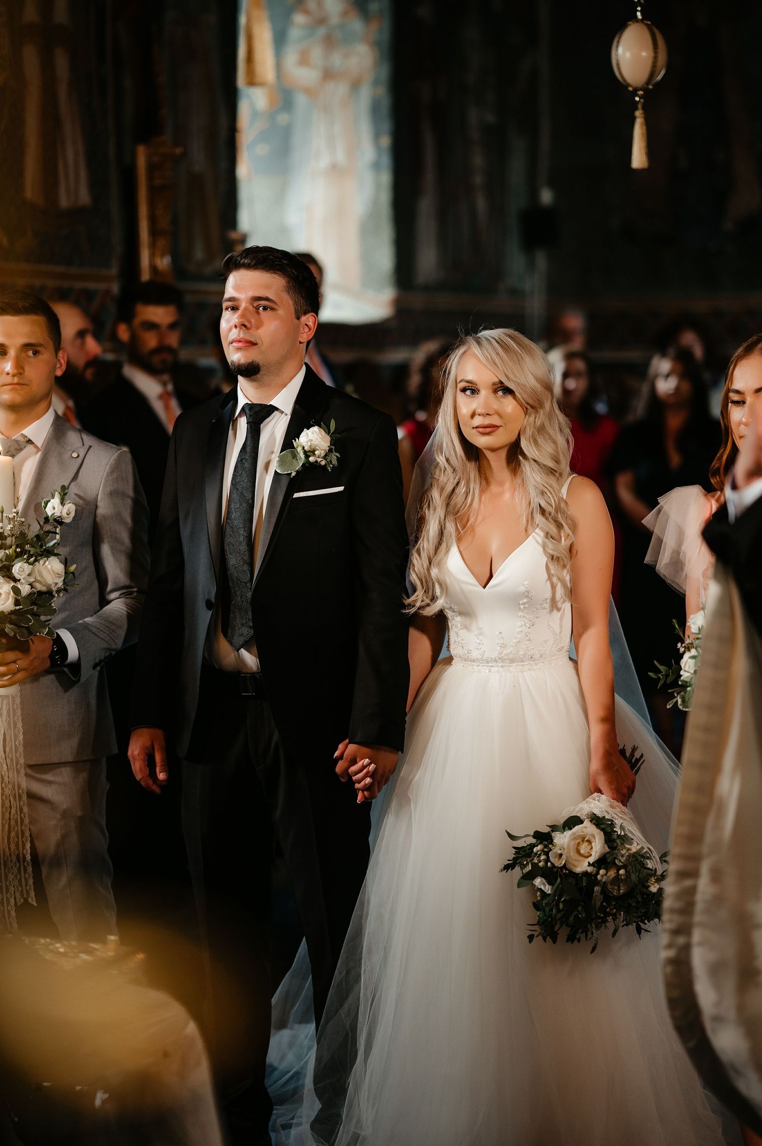 Bride in white tulle gown and groom in black suit holding hands during Orthodox wedding ceremony.