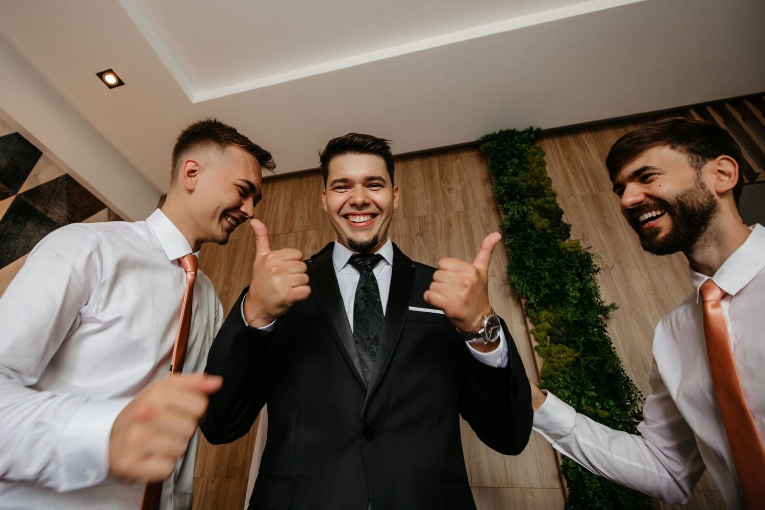 Groom in black tuxedo gives thumbs up with two smiling groomsmen in white shirts before wedding.