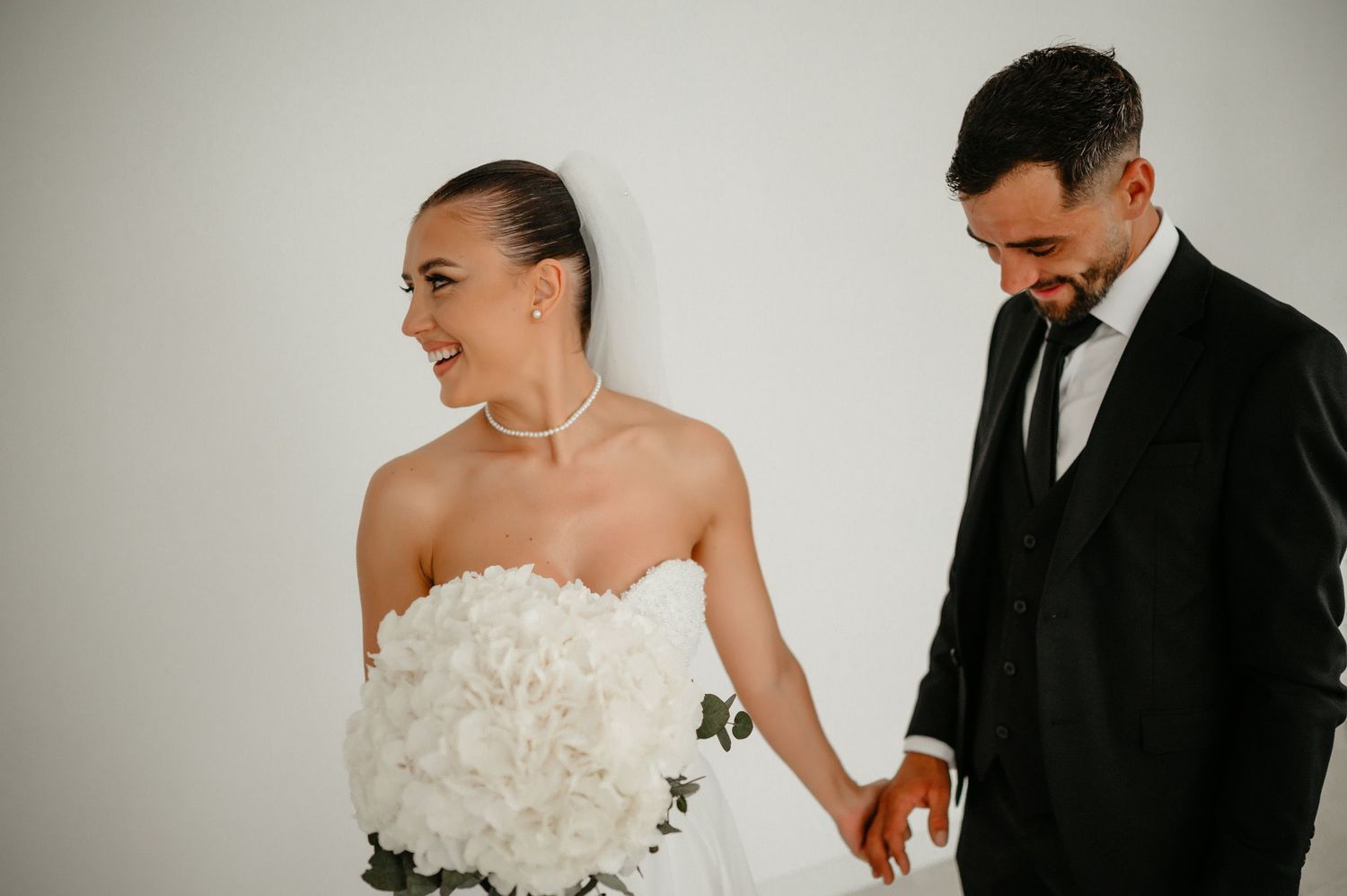 Joyful bride in strapless floral gown holds white bouquet while groom in black suit smiles beside her.