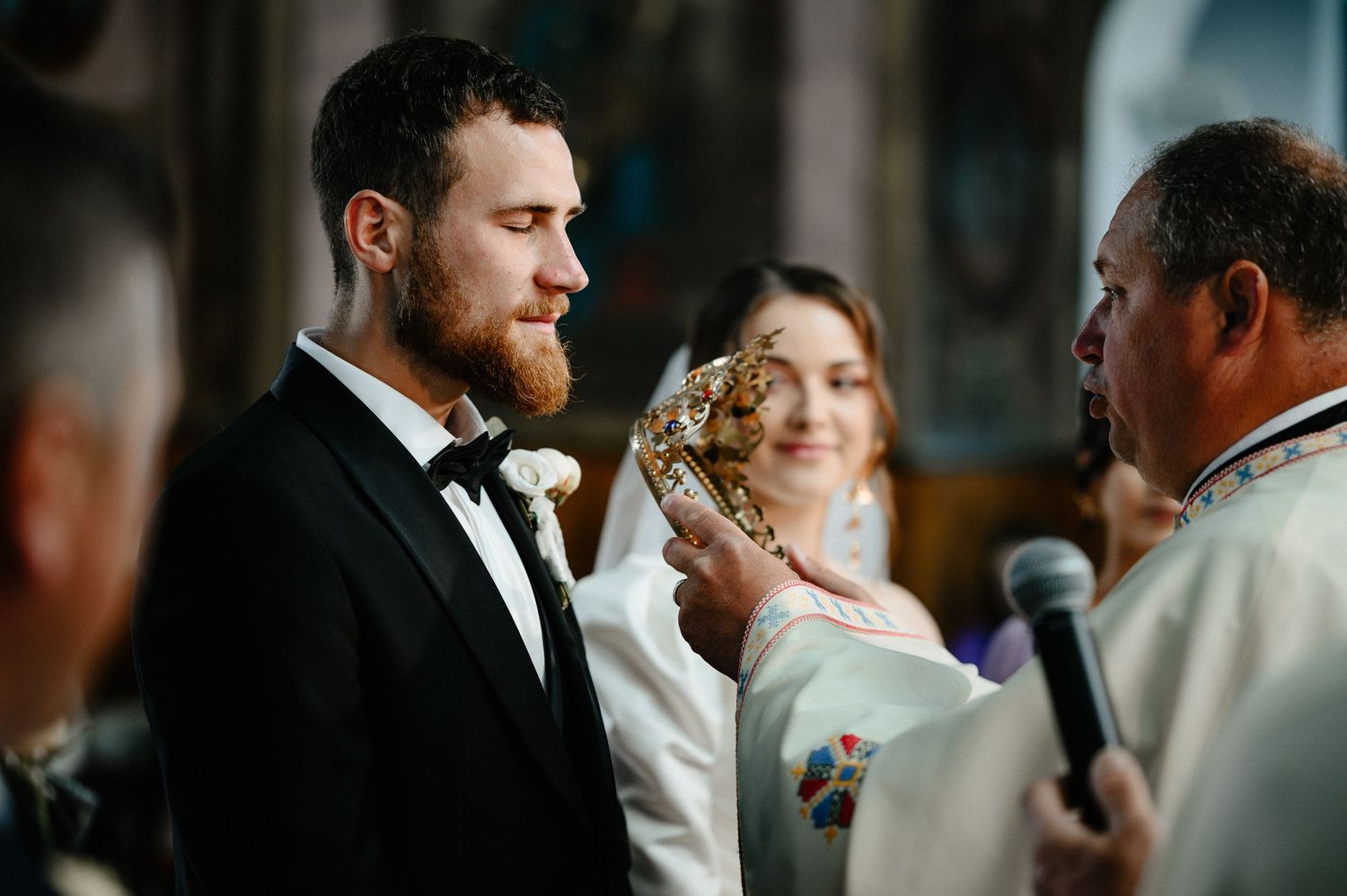 Groom in tuxedo receiving blessing from priest during Orthodox wedding ceremony.