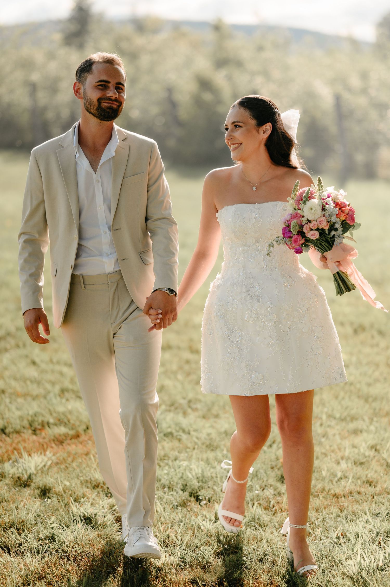 Bride in mini lace dress holding pink bouquet walks hand in hand with groom in beige suit.