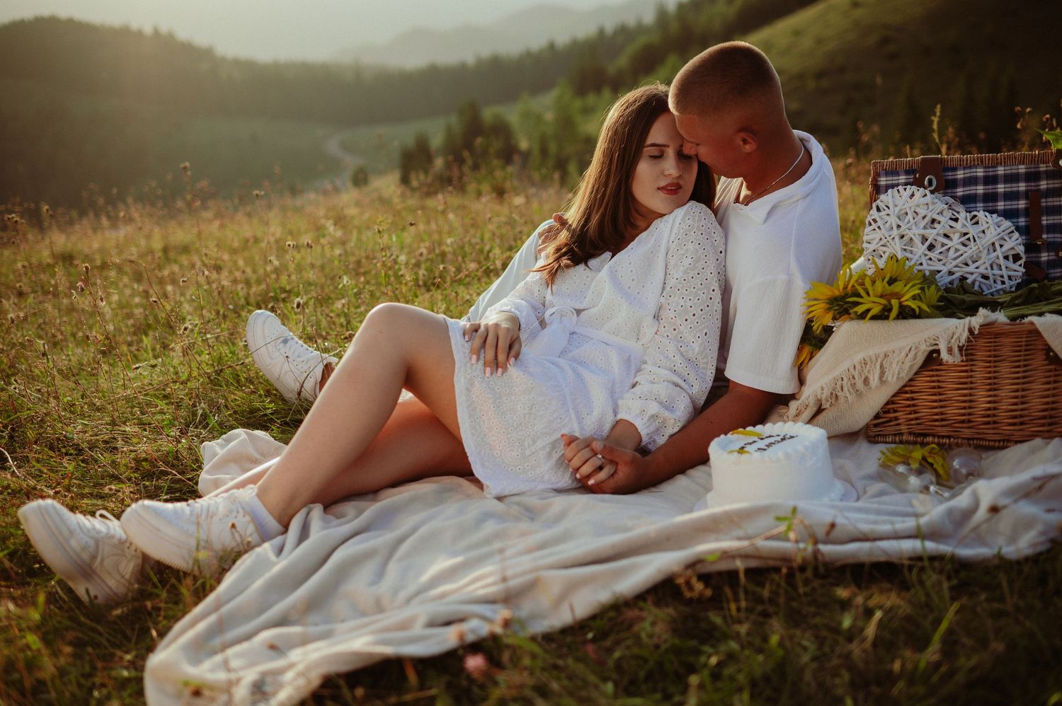 Loving couple cuddling on white blanket with wicker basket in scenic mountain field.