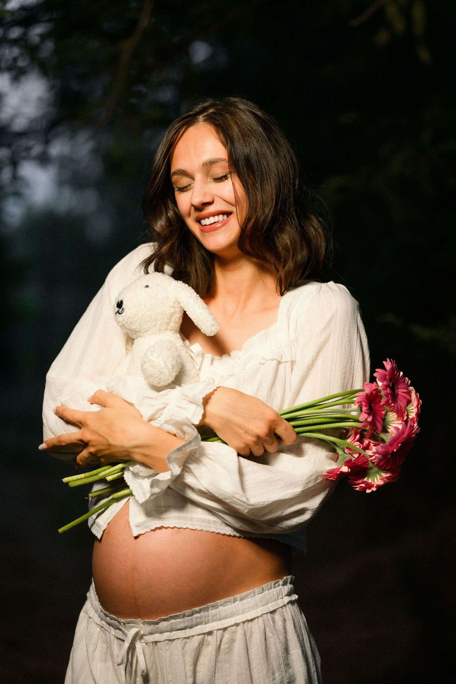 Smiling pregnant woman holding a stuffed bunny and pink gerbera flowers outdoors in soft natural light.