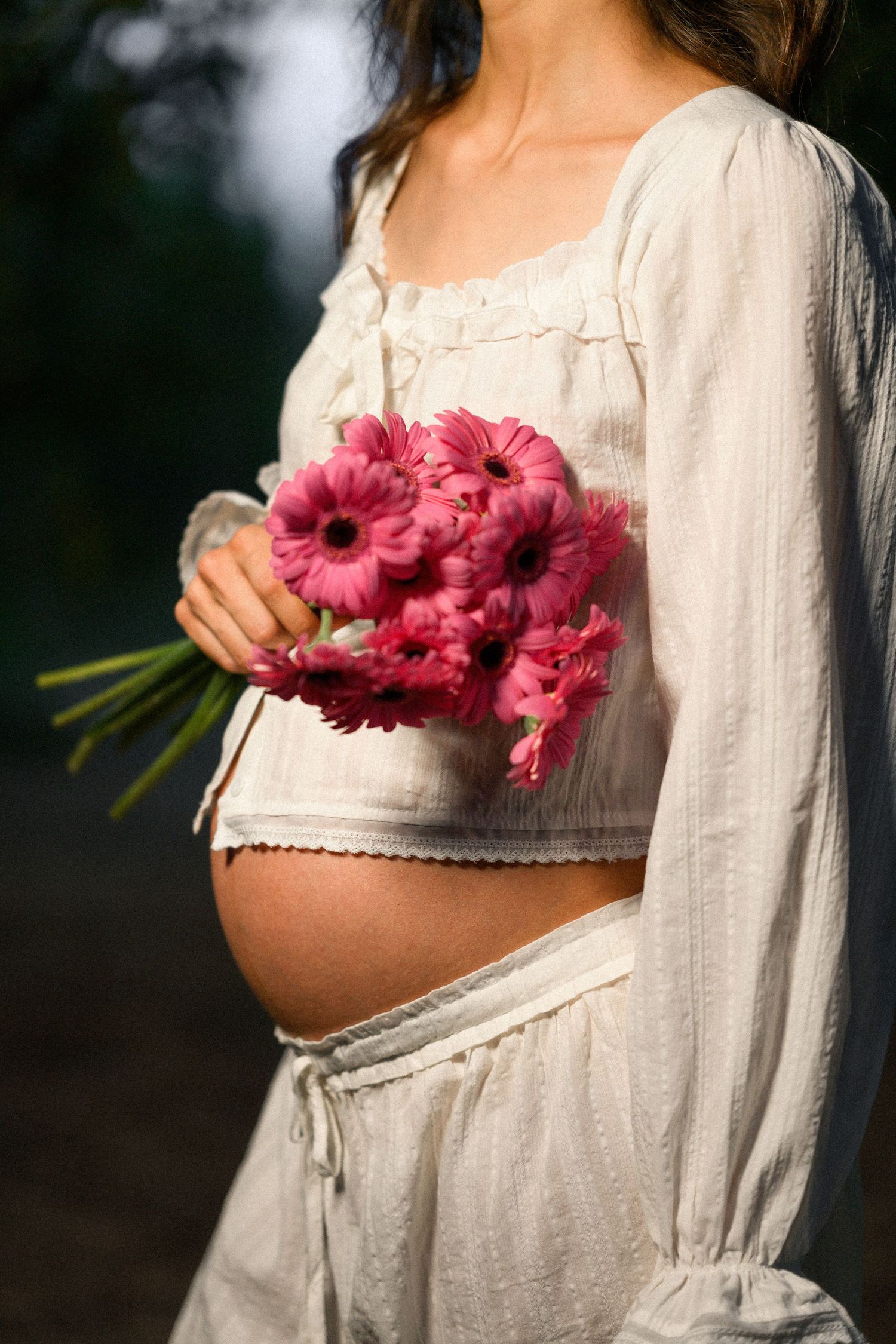 Pregnant woman in white crop top holding pink gerbera daisy bouquet against her bare baby bump outdoors.