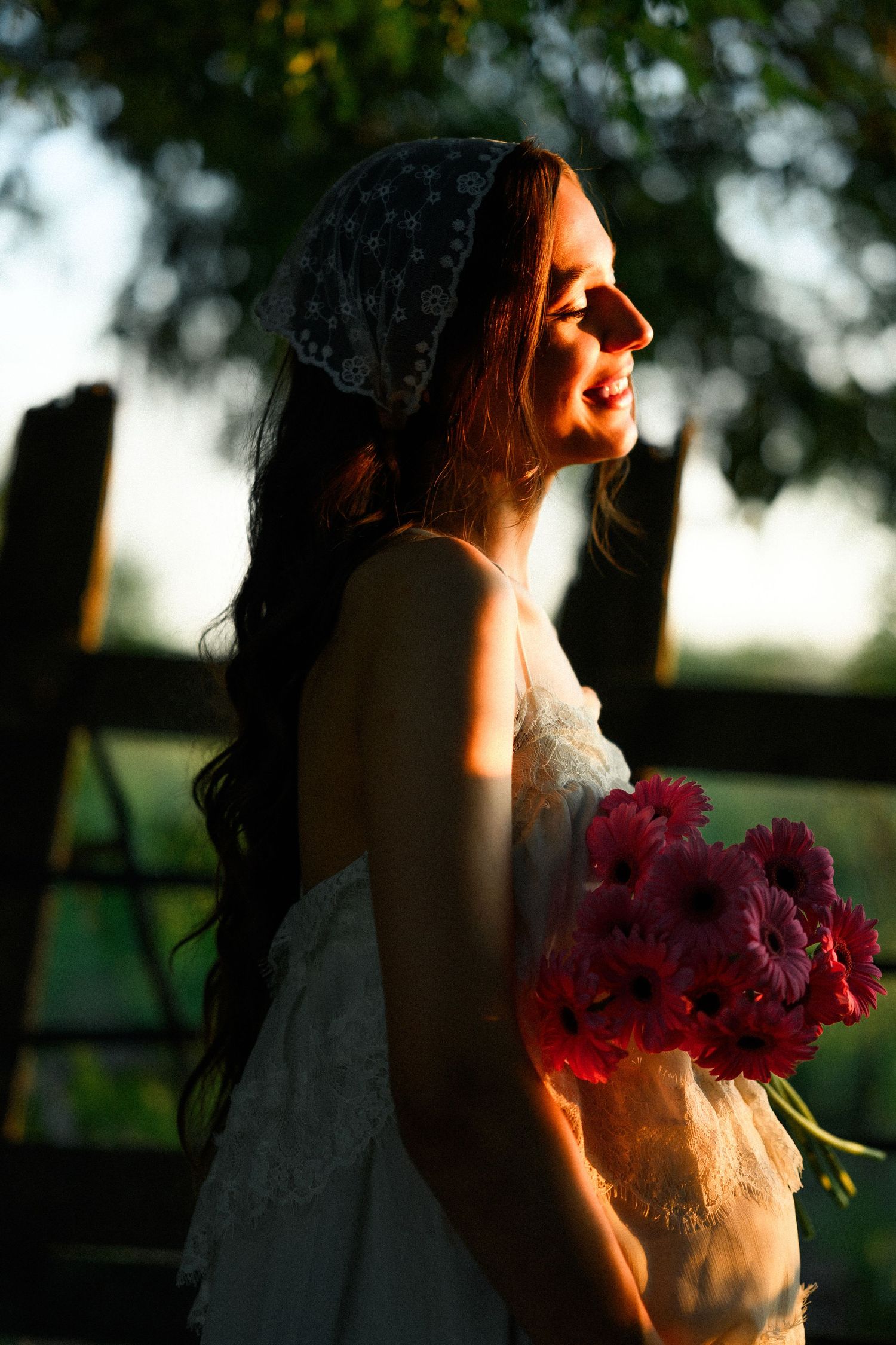 Bride in white lace dress holding pink gerbera bouquet, bathed in golden sunset light outdoors.