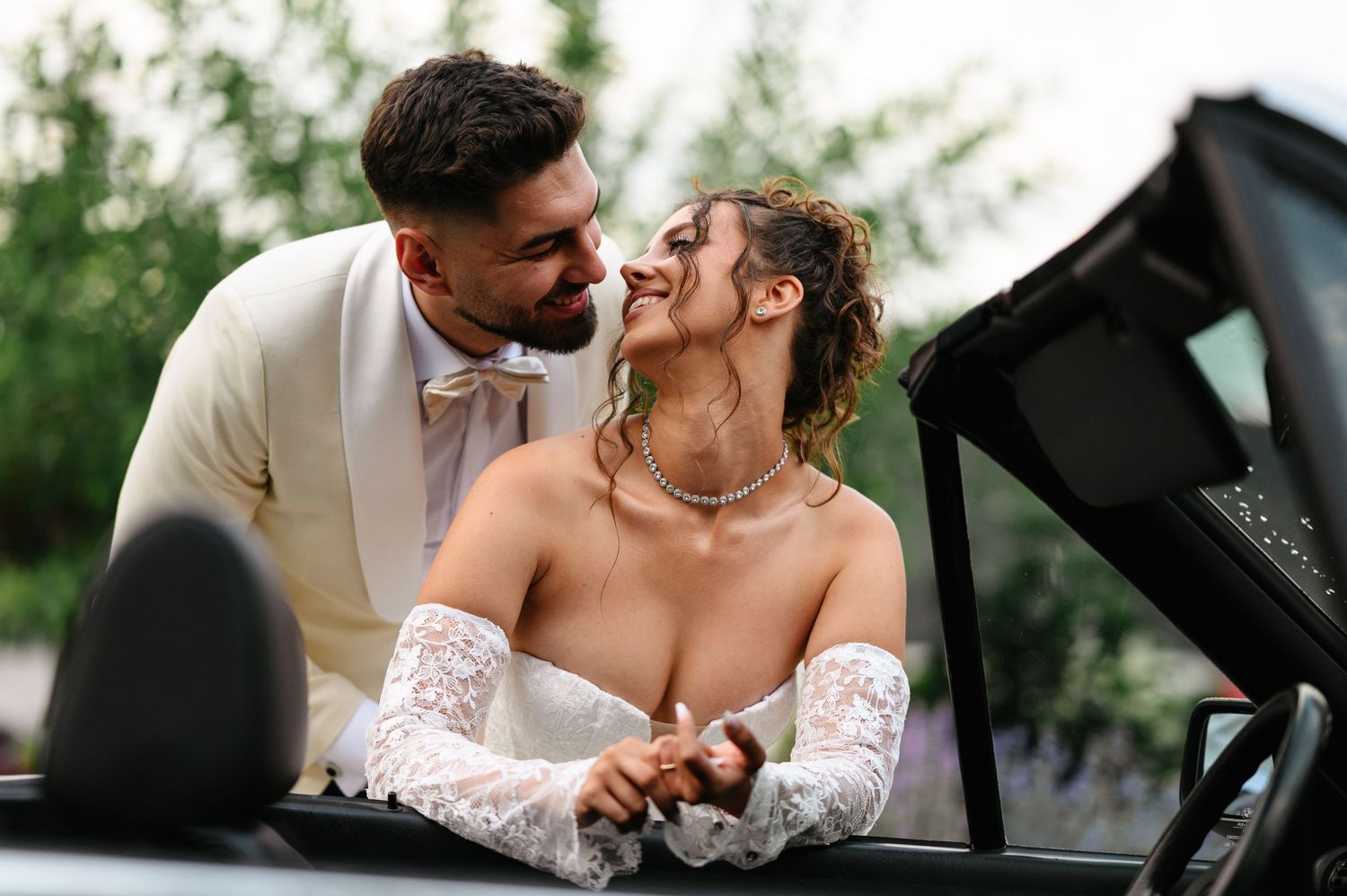 Happy bride and groom sharing a romantic moment in a convertible car, wearing elegant white wedding attire outdoors.