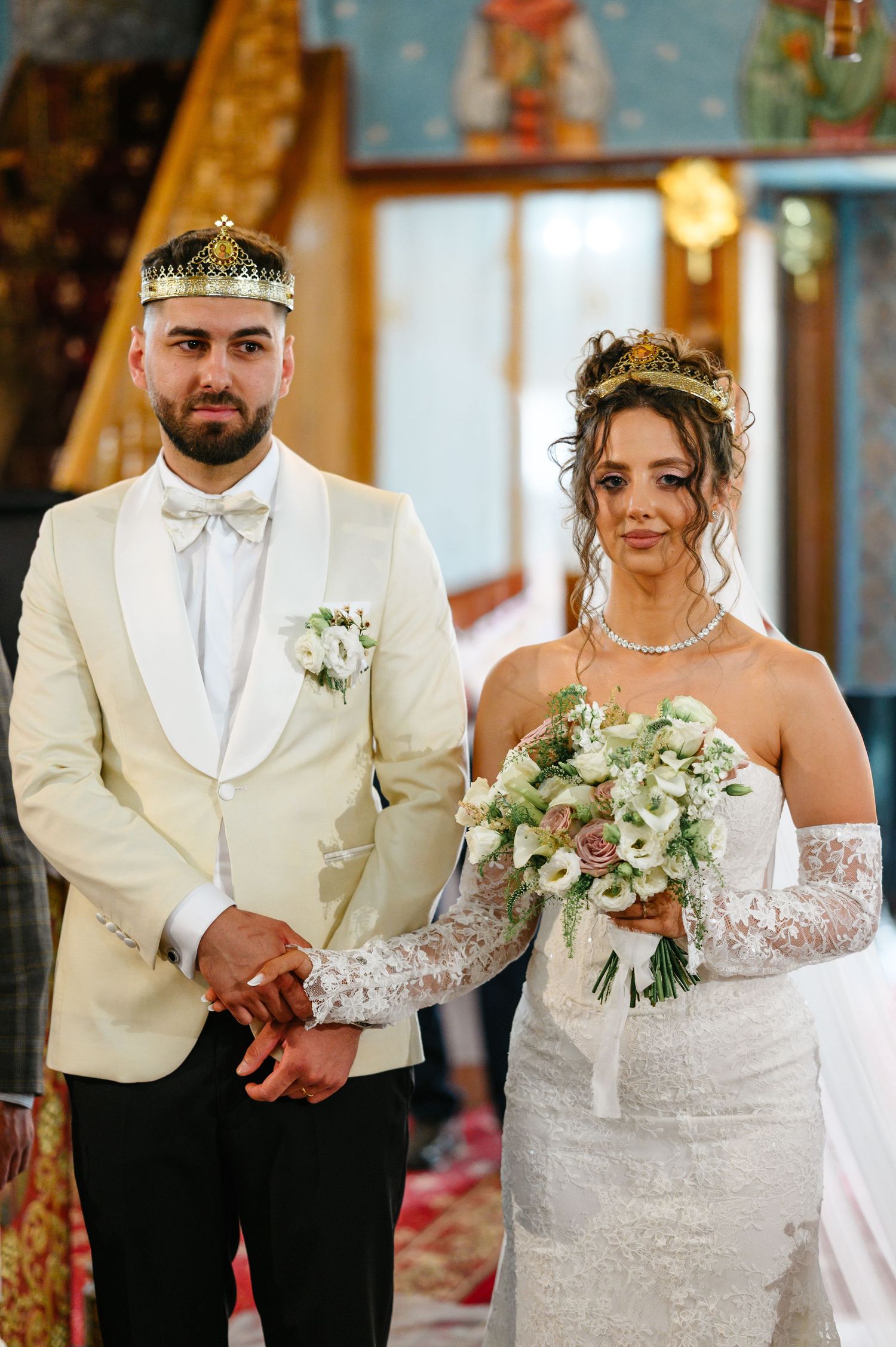 Bride and groom in crowns at Orthodox wedding ceremony, bride holding white floral bouquet, groom in cream tuxedo.