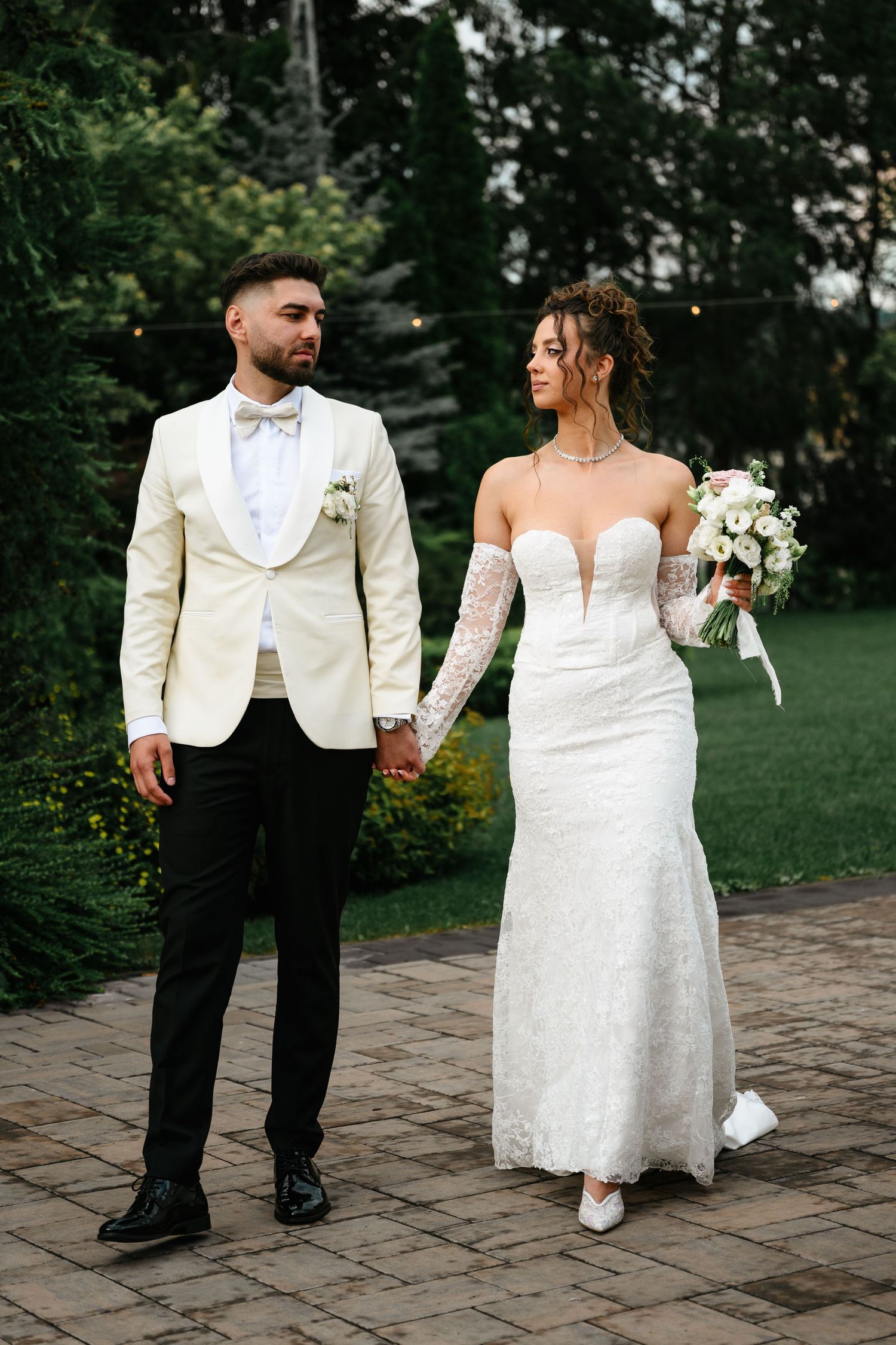 Bride and groom walk hand-in-hand outdoors, she in lace gown with bouquet, he in cream tuxedo jacket.
