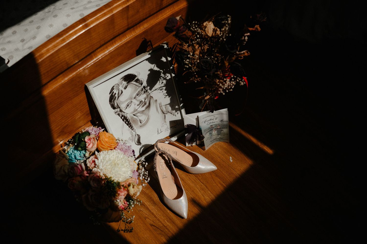 Wedding flat lay with bridal bouquet, heels, framed photo, and floral arrangement on wooden surface in dramatic lighting.