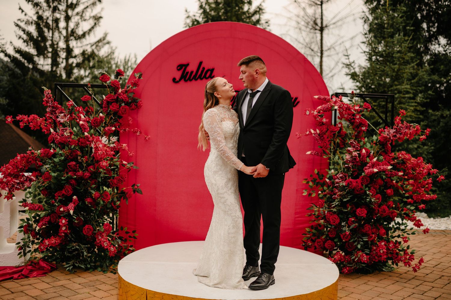 Bride and groom posing before red floral arch with Julia sign.