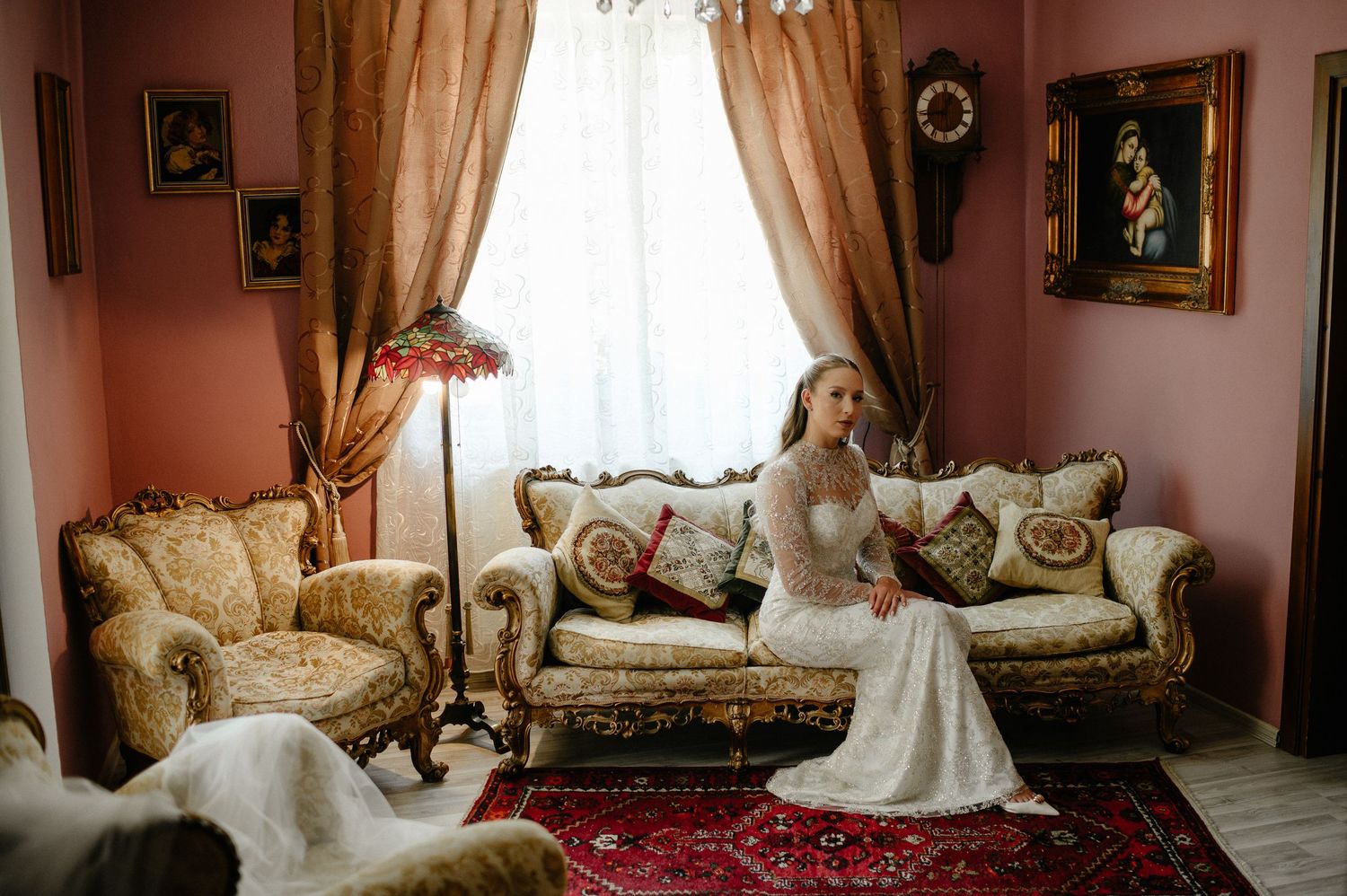 Elegant bride in lace wedding dress poses on golden vintage sofa in ornate pink room.