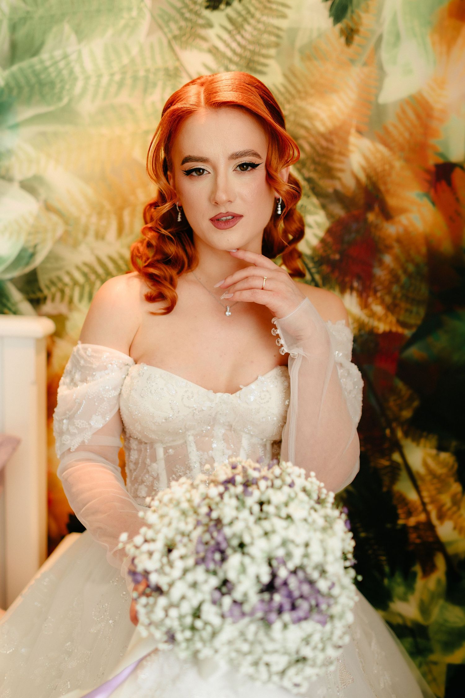Red-haired bride in white lace off-shoulder gown holds baby's breath bouquet before floral backdrop.