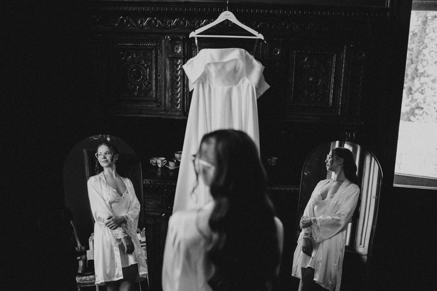 Black and white photo of bride viewing her wedding dress hanging in an ornate dark room.