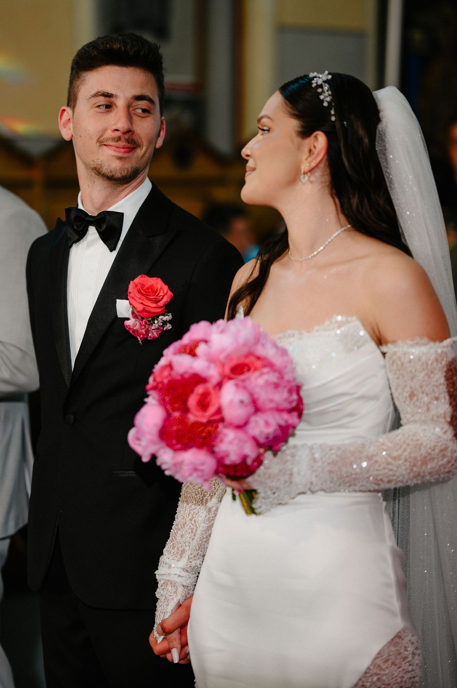 Smiling bride in off-shoulder gown and groom in tuxedo share tender moment during church ceremony.