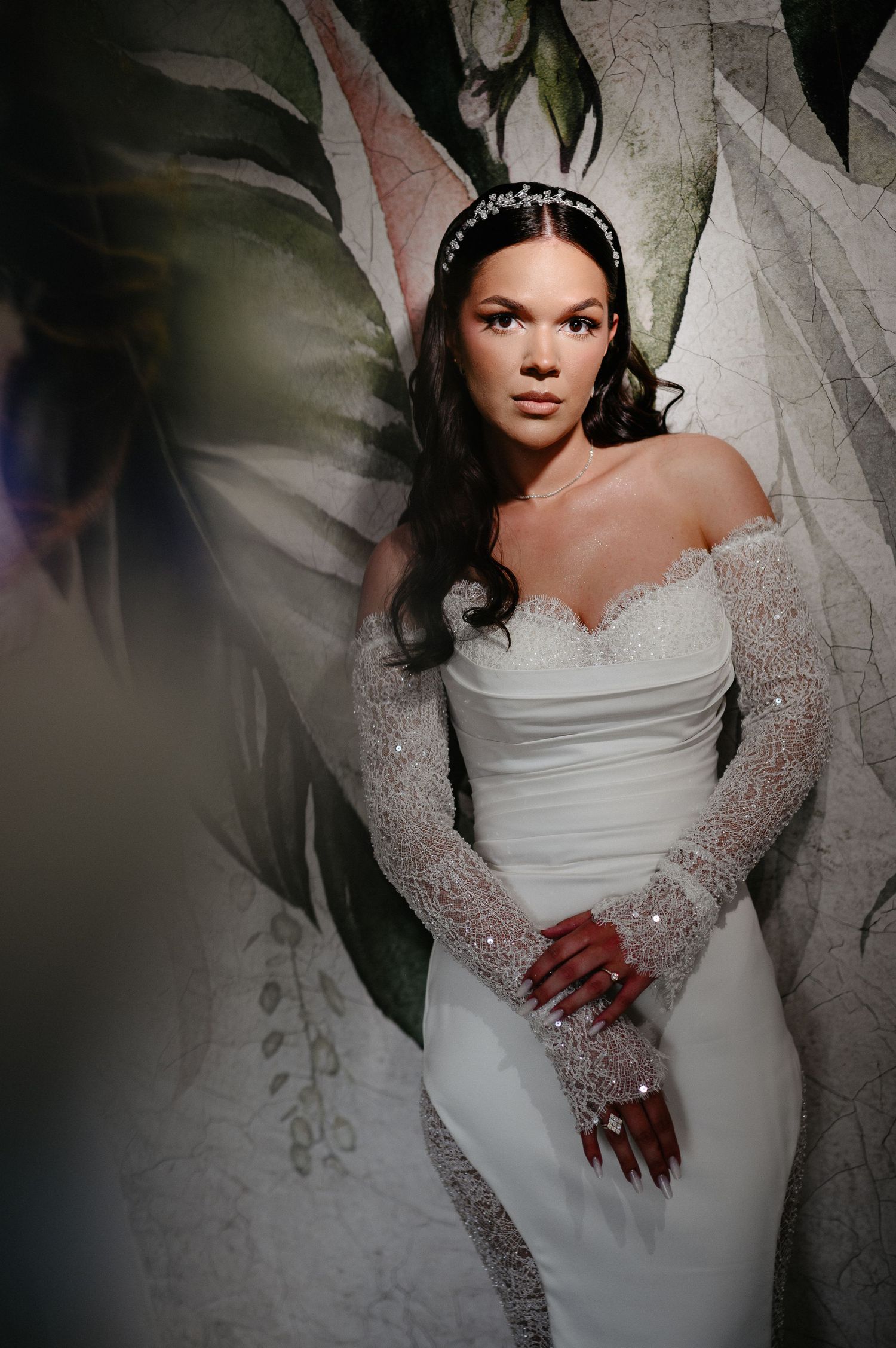 Beautiful bride wearing off-shoulder lace wedding dress and crystal headpiece posing against botanical mural backdrop.