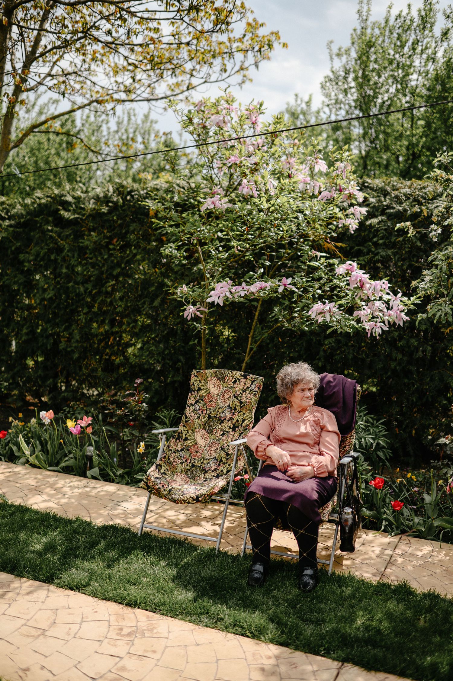 Elderly woman sits peacefully in garden chair surrounded by blooming flowers and lush greenery.