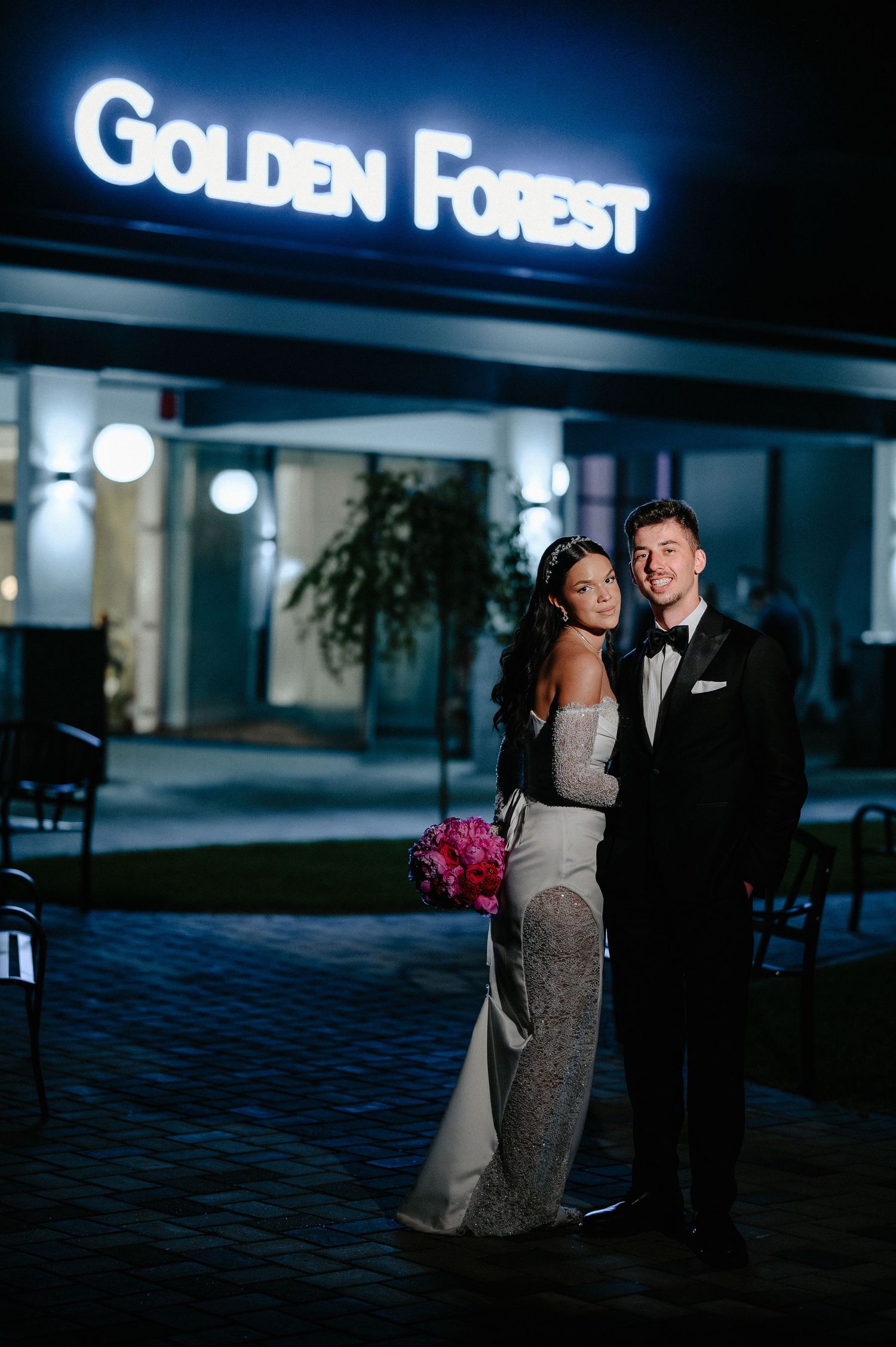 Bride and groom posing together outside illuminated Golden Forest wedding venue sign at night.