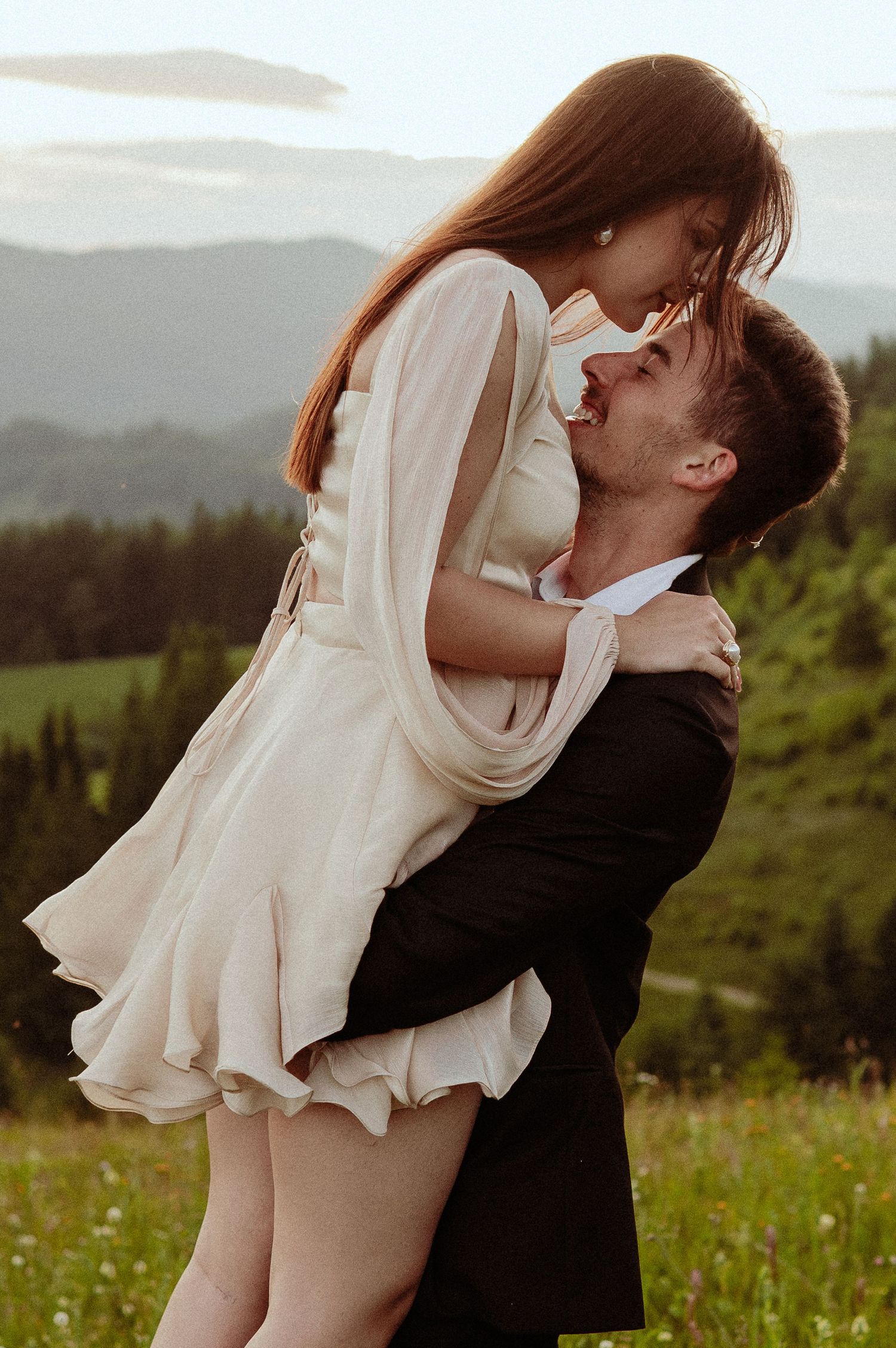 A groom lifts his bride in a flowing cream dress while laughing in a scenic mountain meadow.