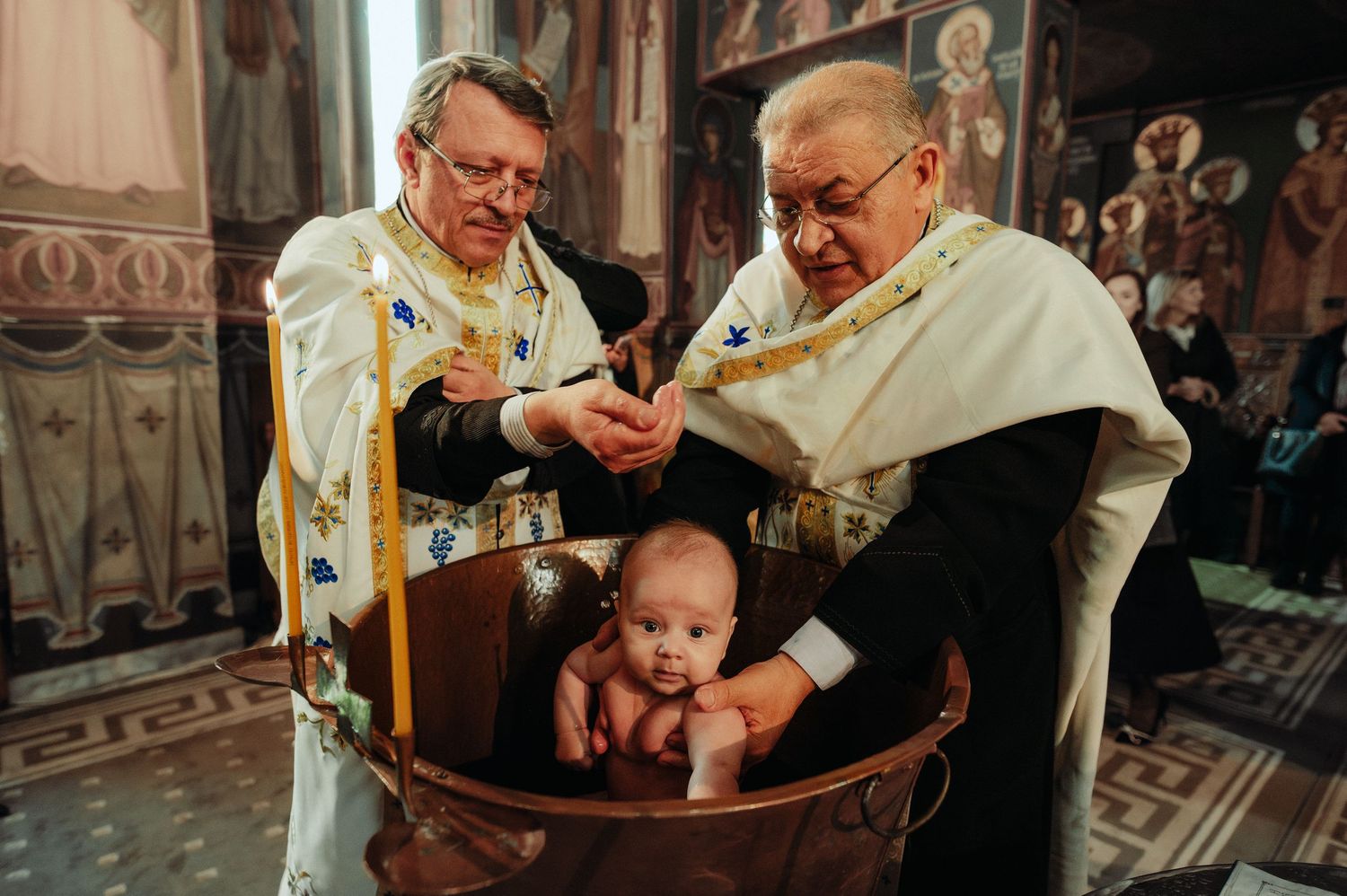 Two Orthodox priests performing a traditional baby baptism in a ceremonial water font.