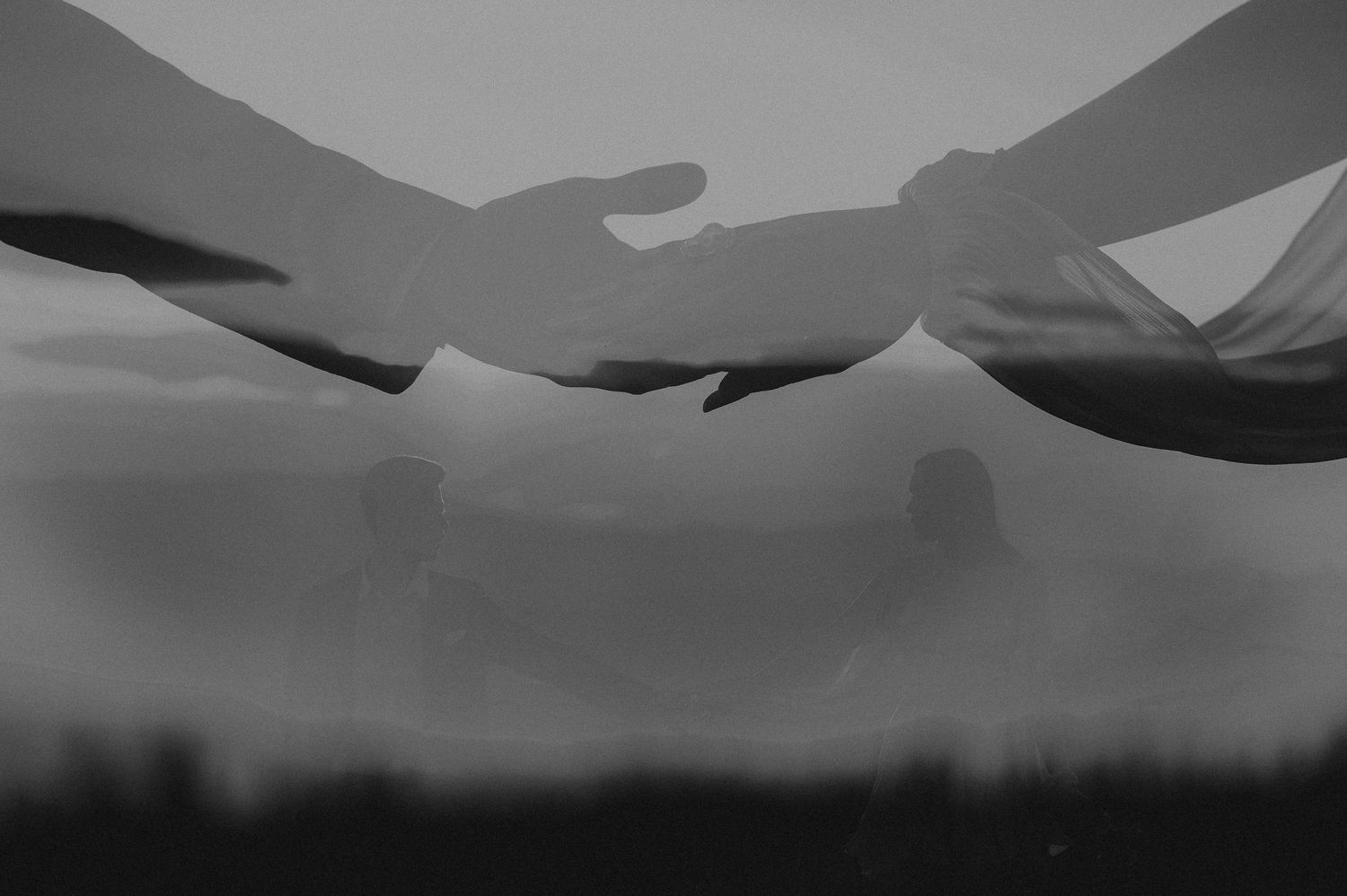 Black and white double exposure of two hands reaching toward each other with a couple silhouette in background.