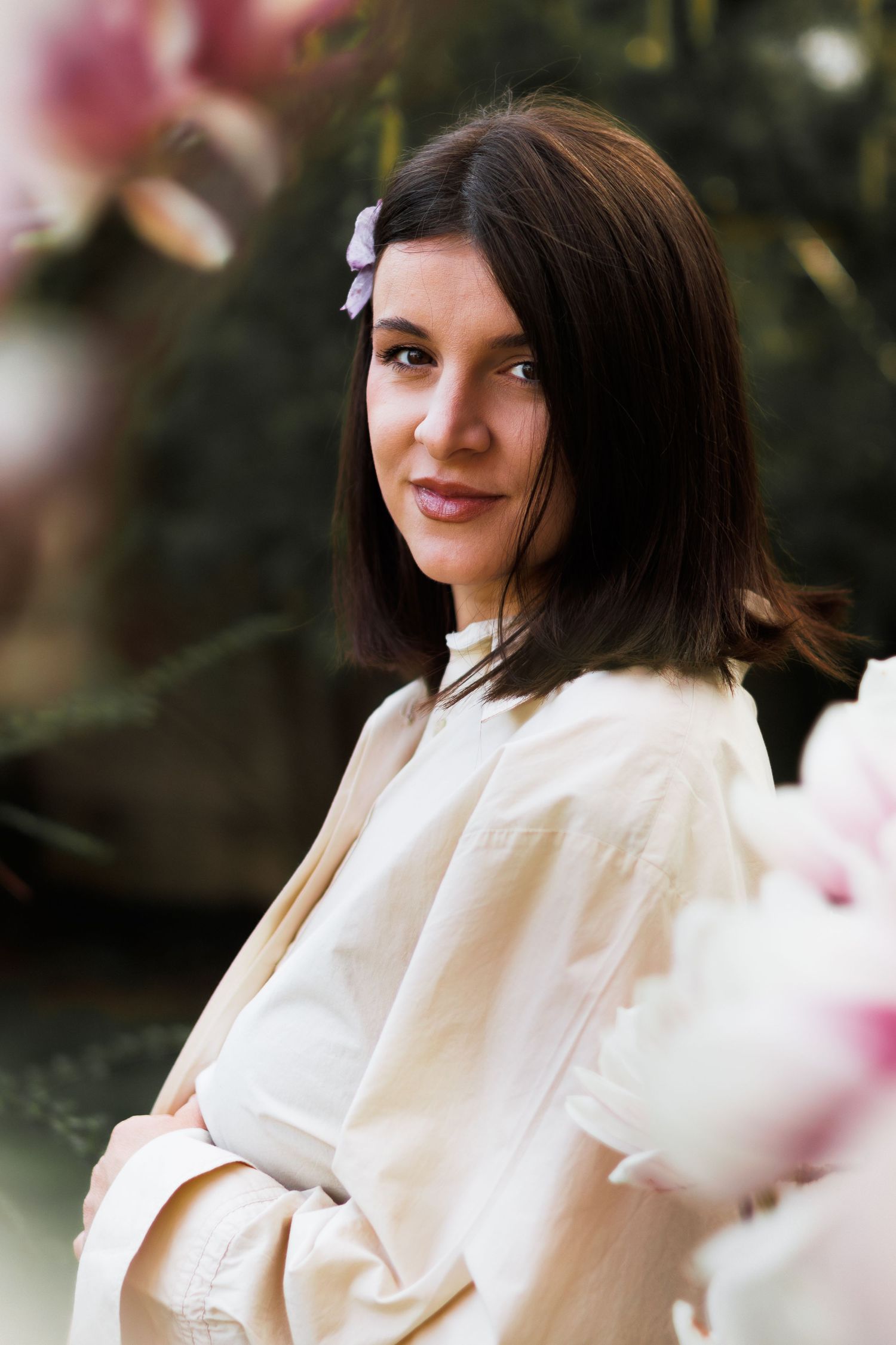 Portrait of dark-haired woman with flower in hair posing near pink magnolia blooms.