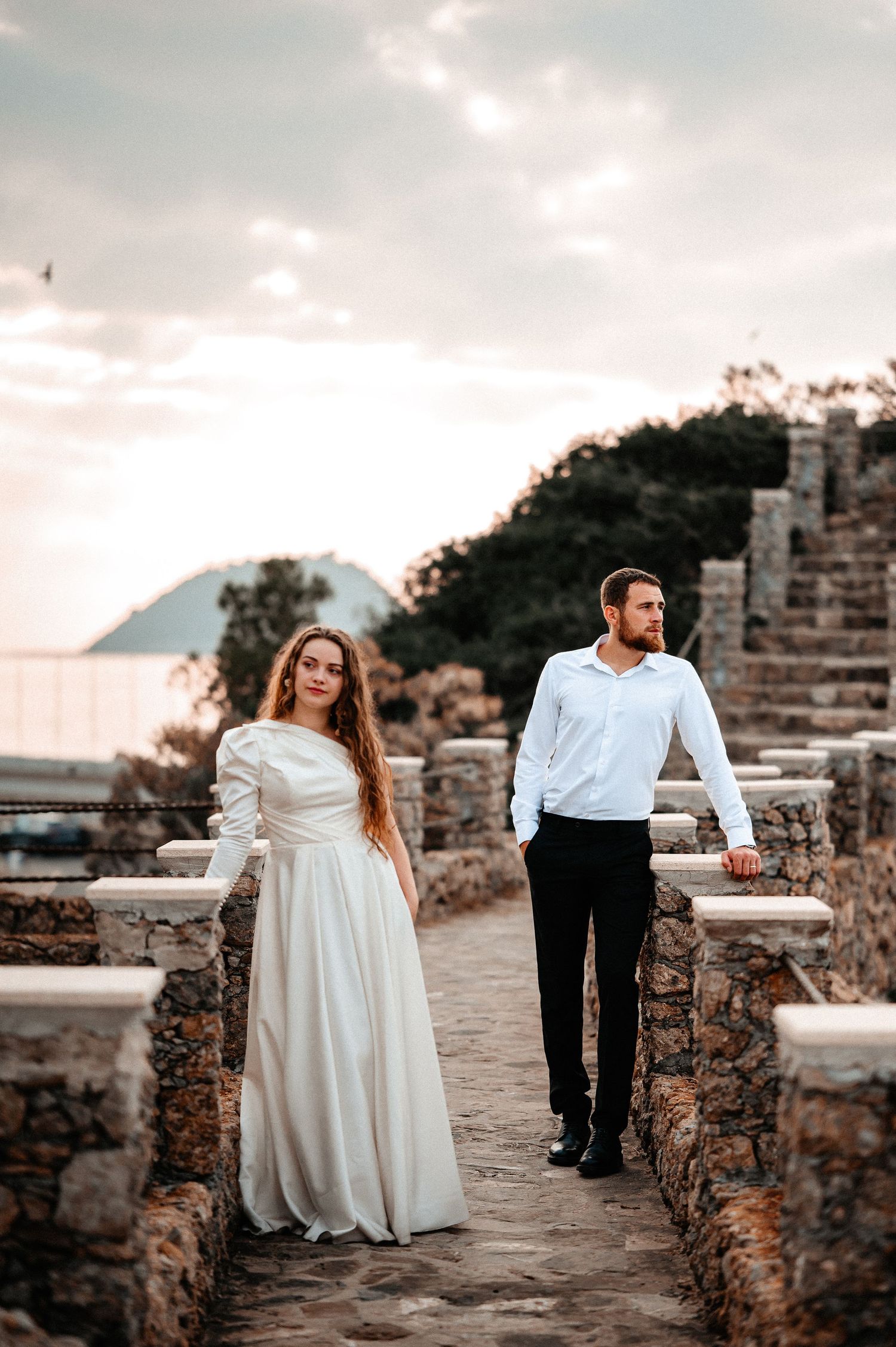 Elegant couple in wedding attire pose on ancient stone steps with sea and mountain backdrop at sunset.