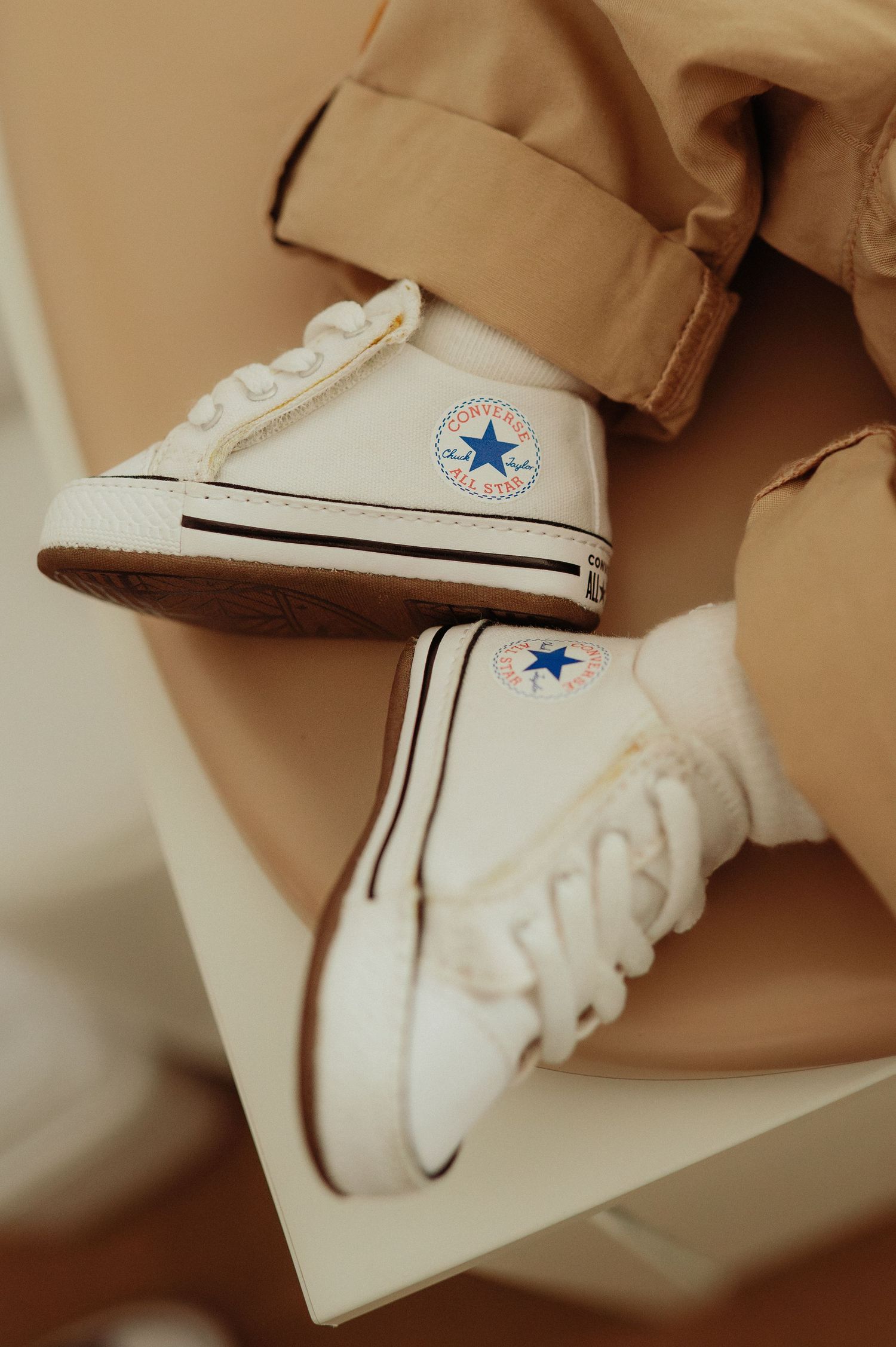 Tiny baby Converse Chuck Taylor high-top white sneakers on infant feet with beige pants.