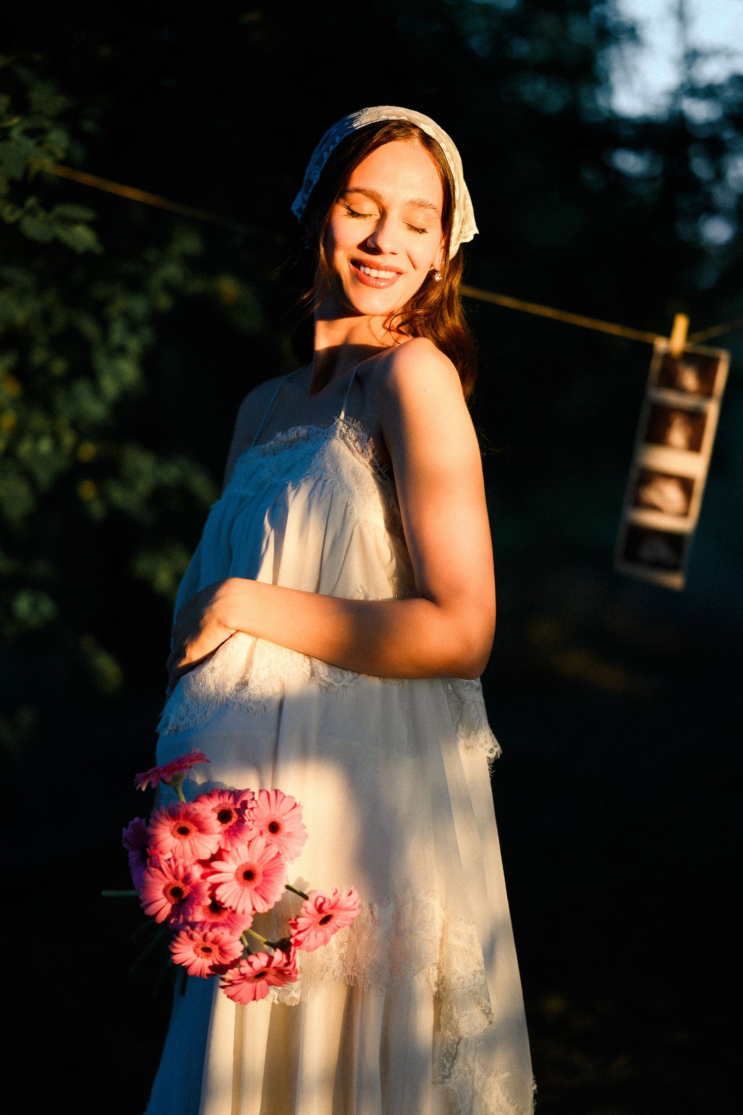 Smiling pregnant woman in white dress holding pink gerbera flowers outdoors in golden sunset light.