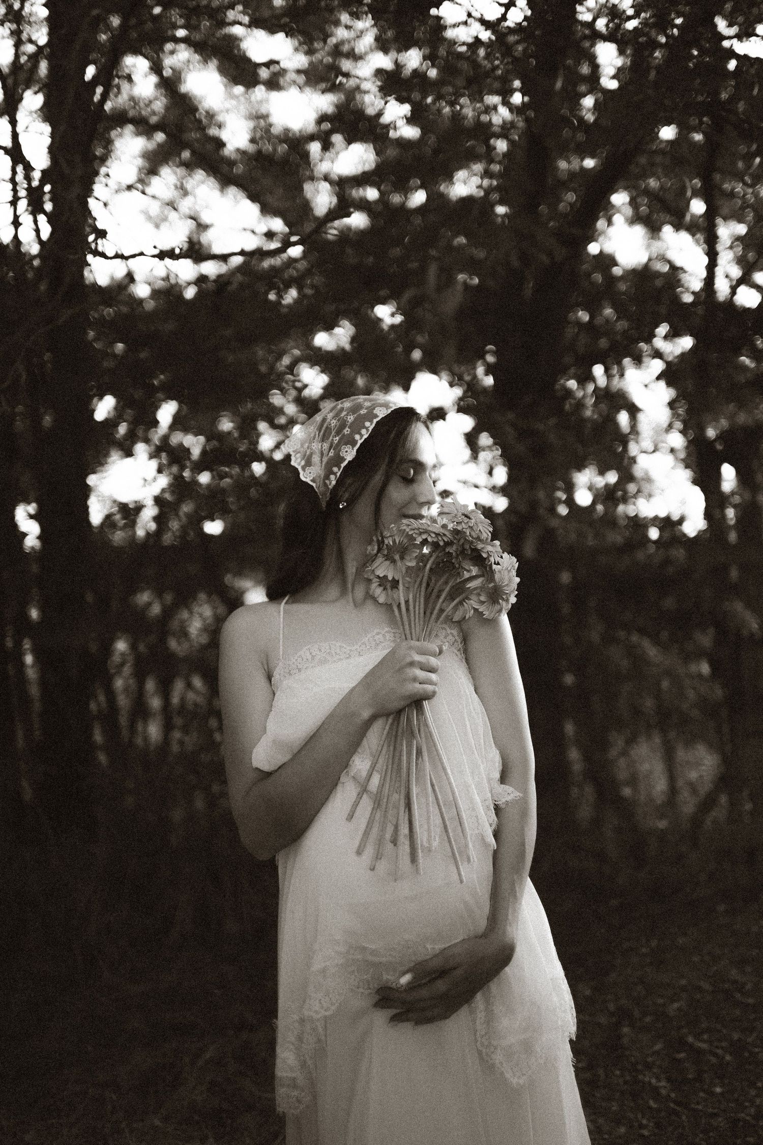 Pregnant woman in white lace dress holding wildflowers in a forest, black and white maternity photo.