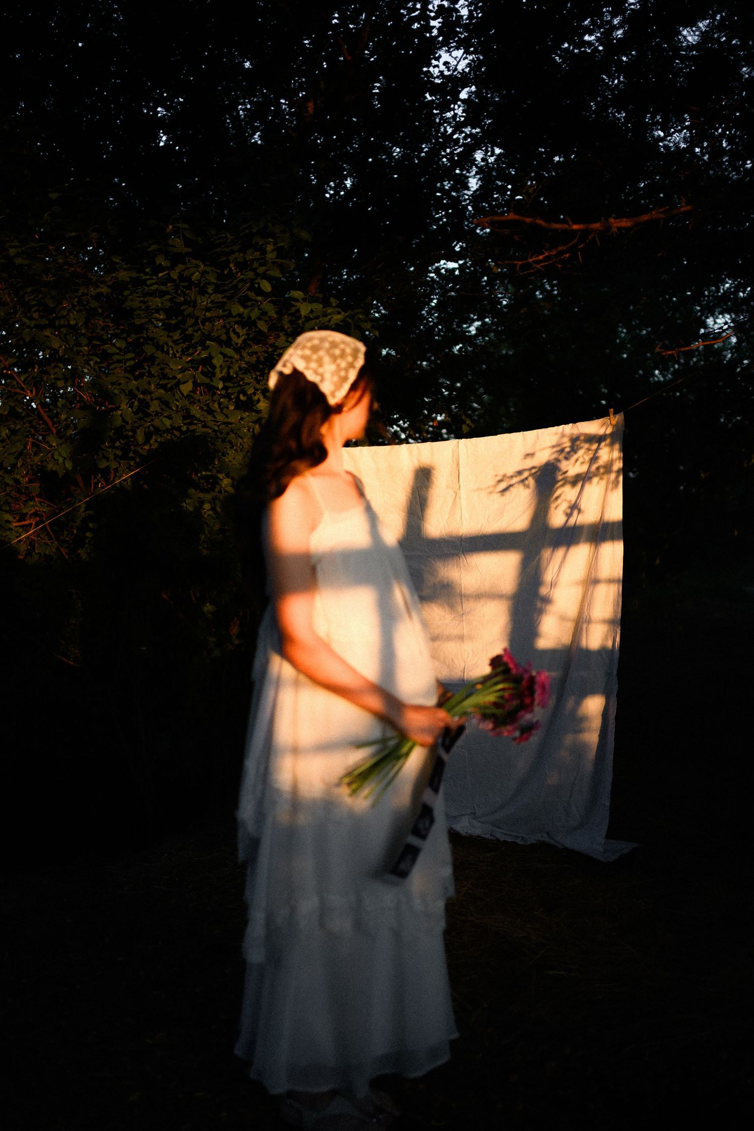 Woman in white dress holding flower bouquet standing by hanging sheet in golden sunlight among dark trees.